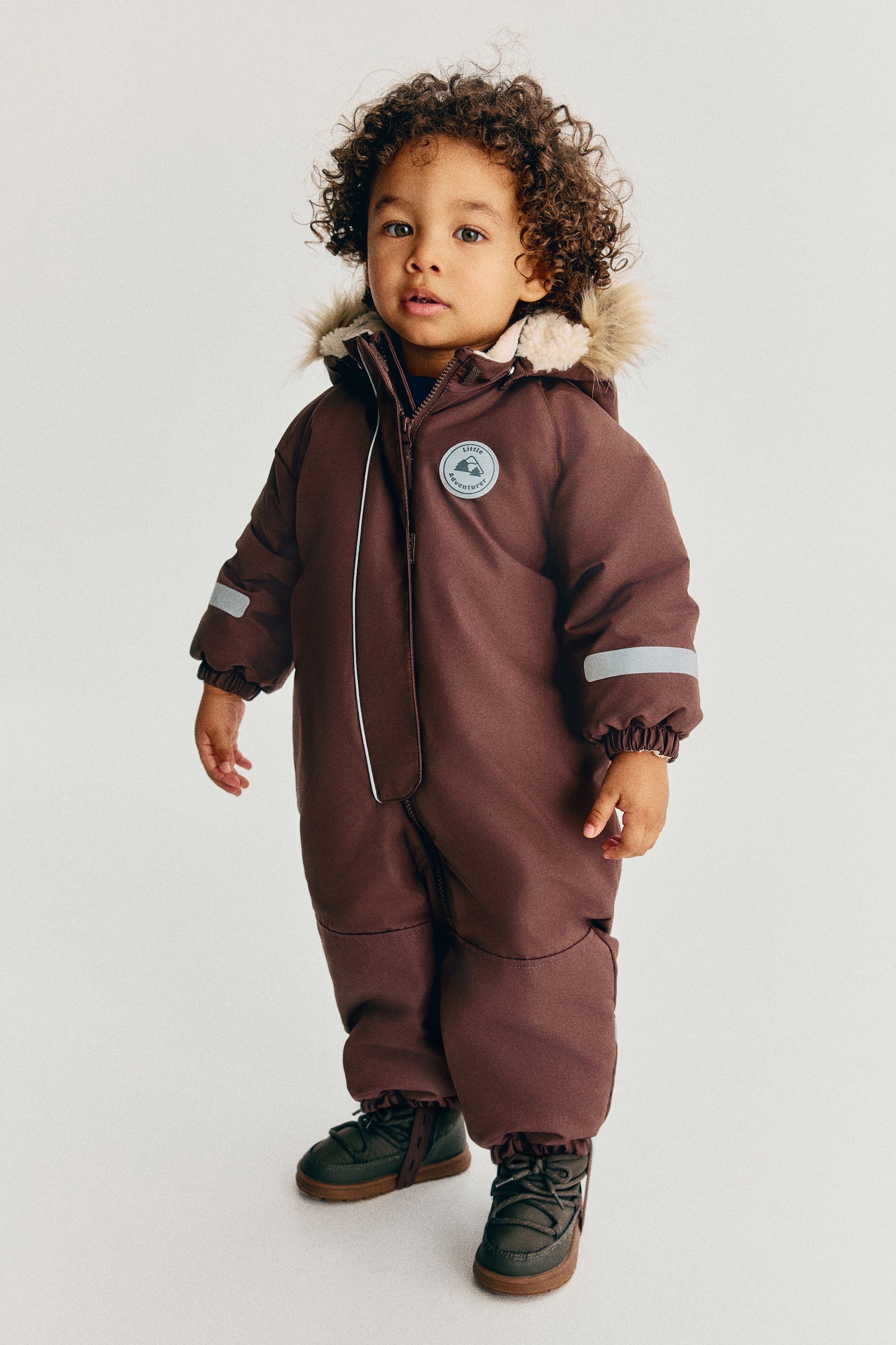 View larger image: A brown one-piece snowsuit is displayed from the front, featuring a hood with a light-coloured faux-fur trim and fleece lining. The snowsuit shows a front zipper closure with reflective piping, a circular chest patch with a mountain design, reflective bands on the sleeves, and elasticized cuffs.