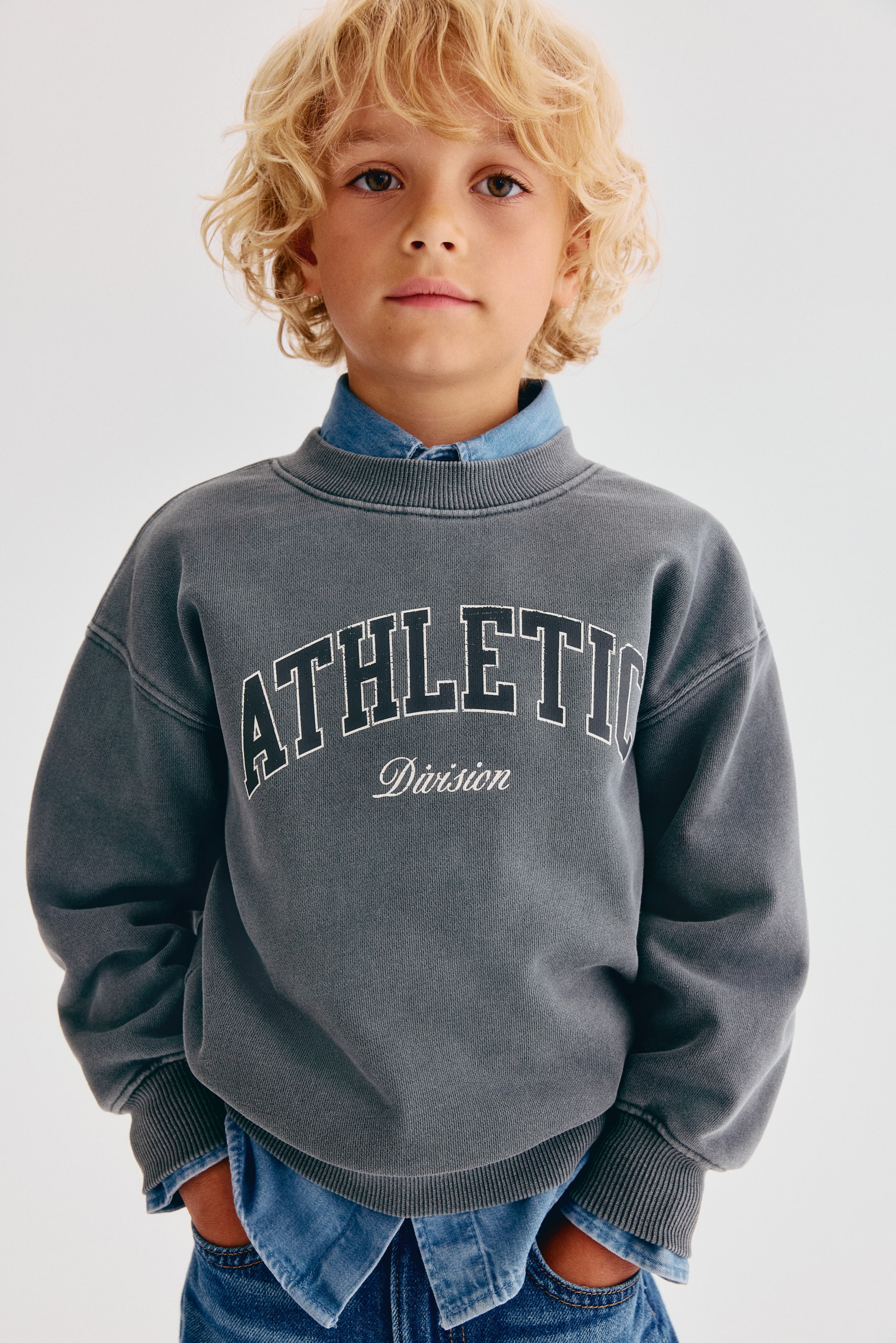 View larger image: Showing from the front, this loose-fit, washed dark grey sweatshirt displays a white graphic design with the arched text "ATHLETIC" above "Division" in script. The sweatshirt features a round, rib-trimmed neckline, dropped shoulders, and ribbed cuffs and hem, and is layered over a light blue denim collared shirt.