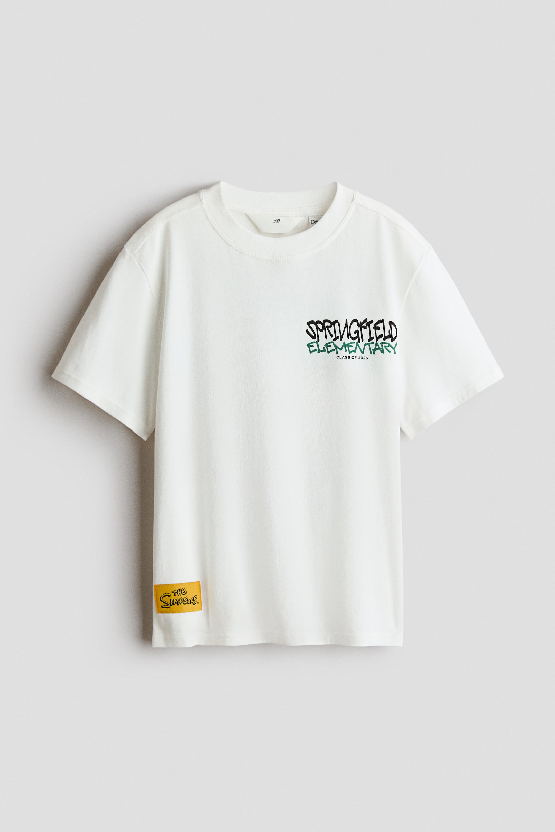 View larger image: A white t-shirt is facing front, featuring a round, ribbed neckline, short sleeves, and a straight-cut hem. A graffiti-style print reading 'SPRINGFIELD ELEMENTARY CLASS OF 2026' is appearing on the front chest, with a small yellow 'The Simpsons' tag on the bottom left hem.
