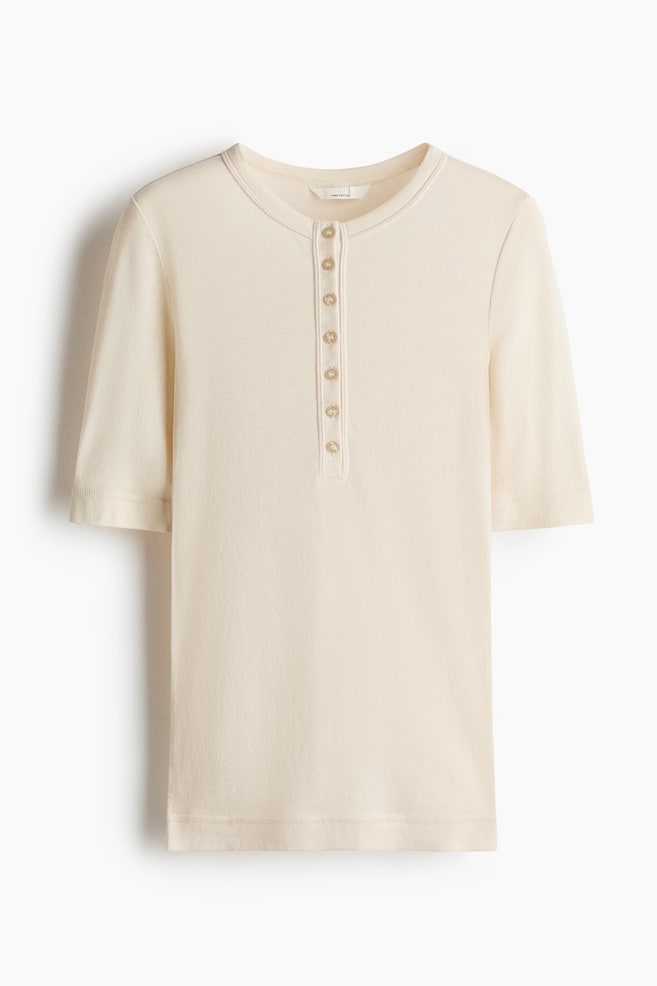 A cream henley shirt is shown from the front, featuring a round neckline with visible stitching and a seven-button placket. It has short sleeves and a straight hem. - 1