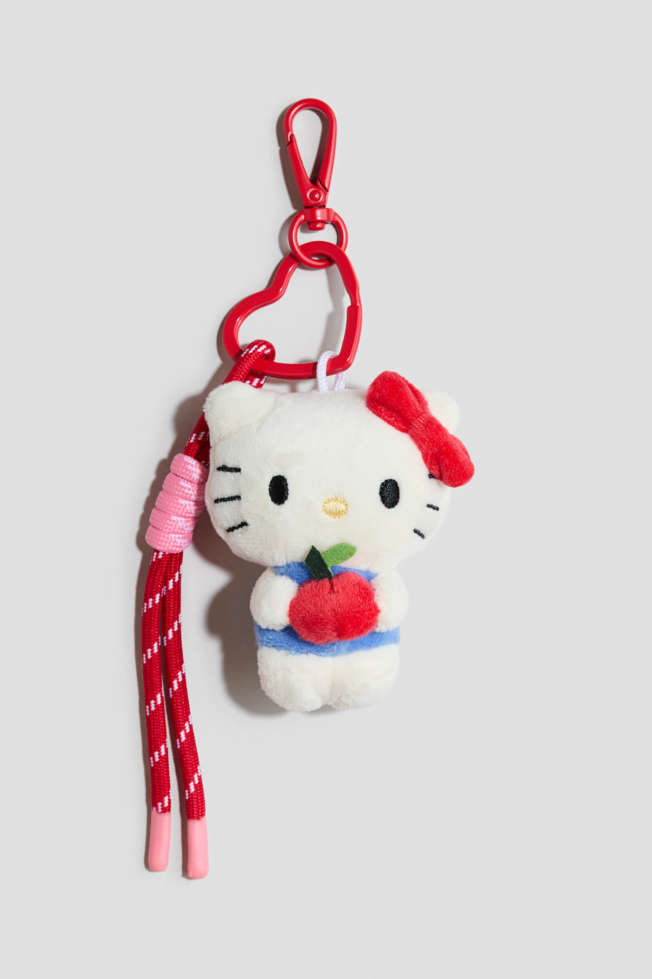 Grotere afbeelding bekijken: Een tassenhanger toont een wit pluchen Hello Kitty-figuurtje, gekleed in een blauw-wit gestreepte outfit en met een rode appel in de hand. Het is bevestigd aan een rood hartvormig ringetje, dat verbonden is met een rode karabijnhaak en ook is voorzien van een rood-wit gestreept koord met roze plastic uiteinden.