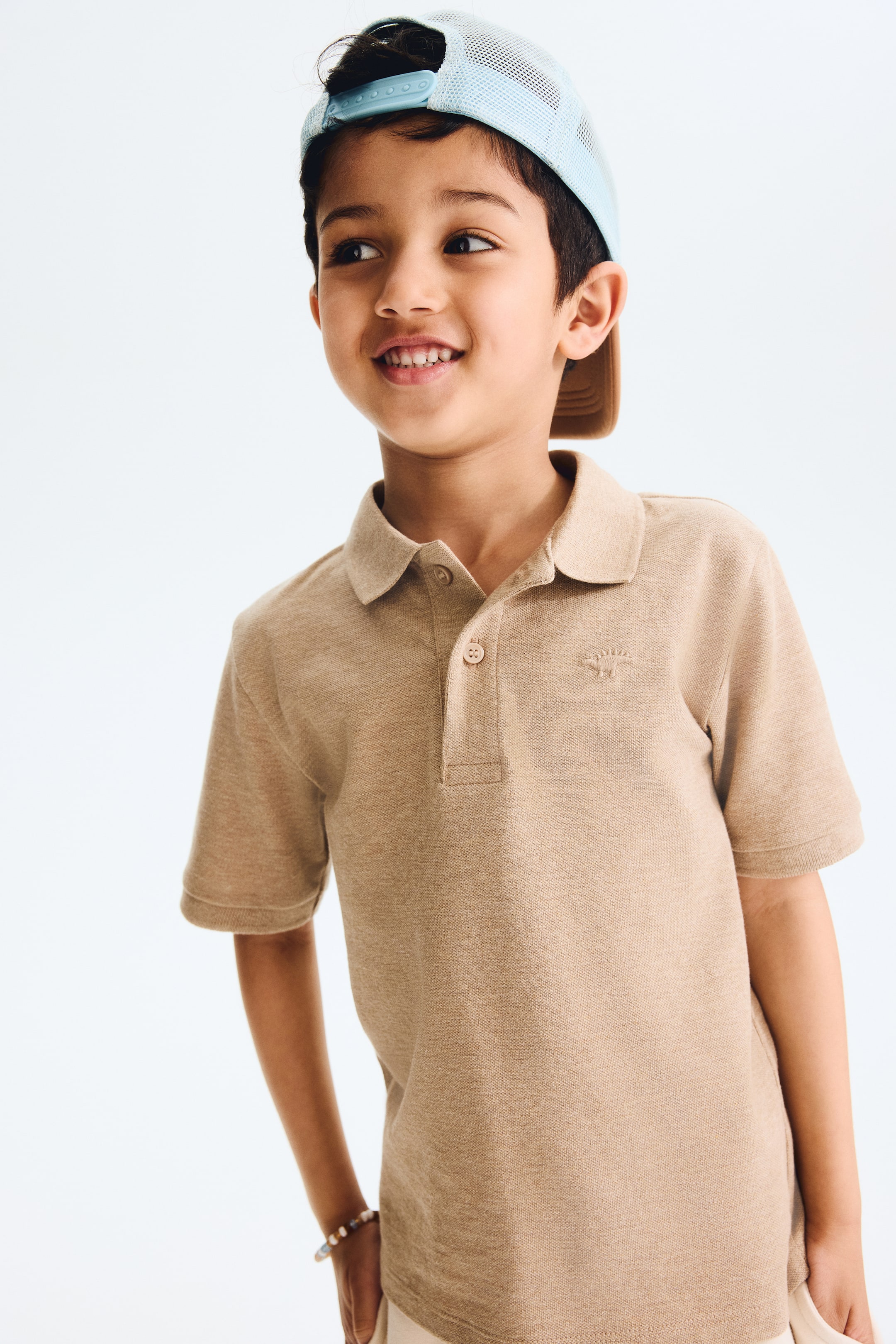 View larger image: Presenting a tan pique knit polo shirt, showcasing a pointed collar, a two-button placket, and a small embroidered dinosaur on the left chest.