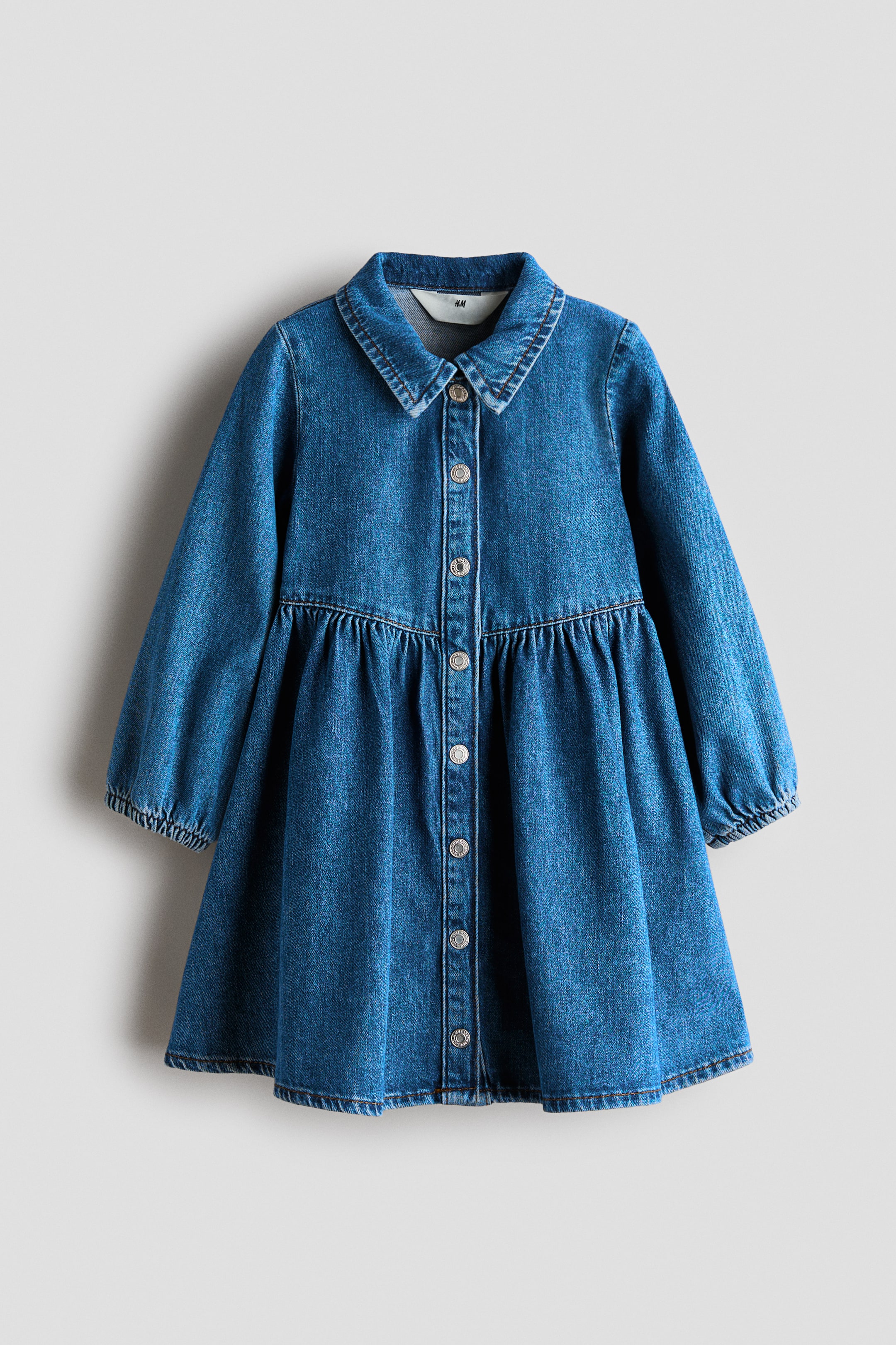 Büyük resmi görüntüle: Önden görünen mavi denim elbise, sivri bir yakaya, metalik düğmeli tam boy düğme patına ve elastik manşetli uzun kollara sahiptir. Elbise, kloş ve akışkan bir eteğe dönüşen büzgülü bir bel dikişi sergiliyor.