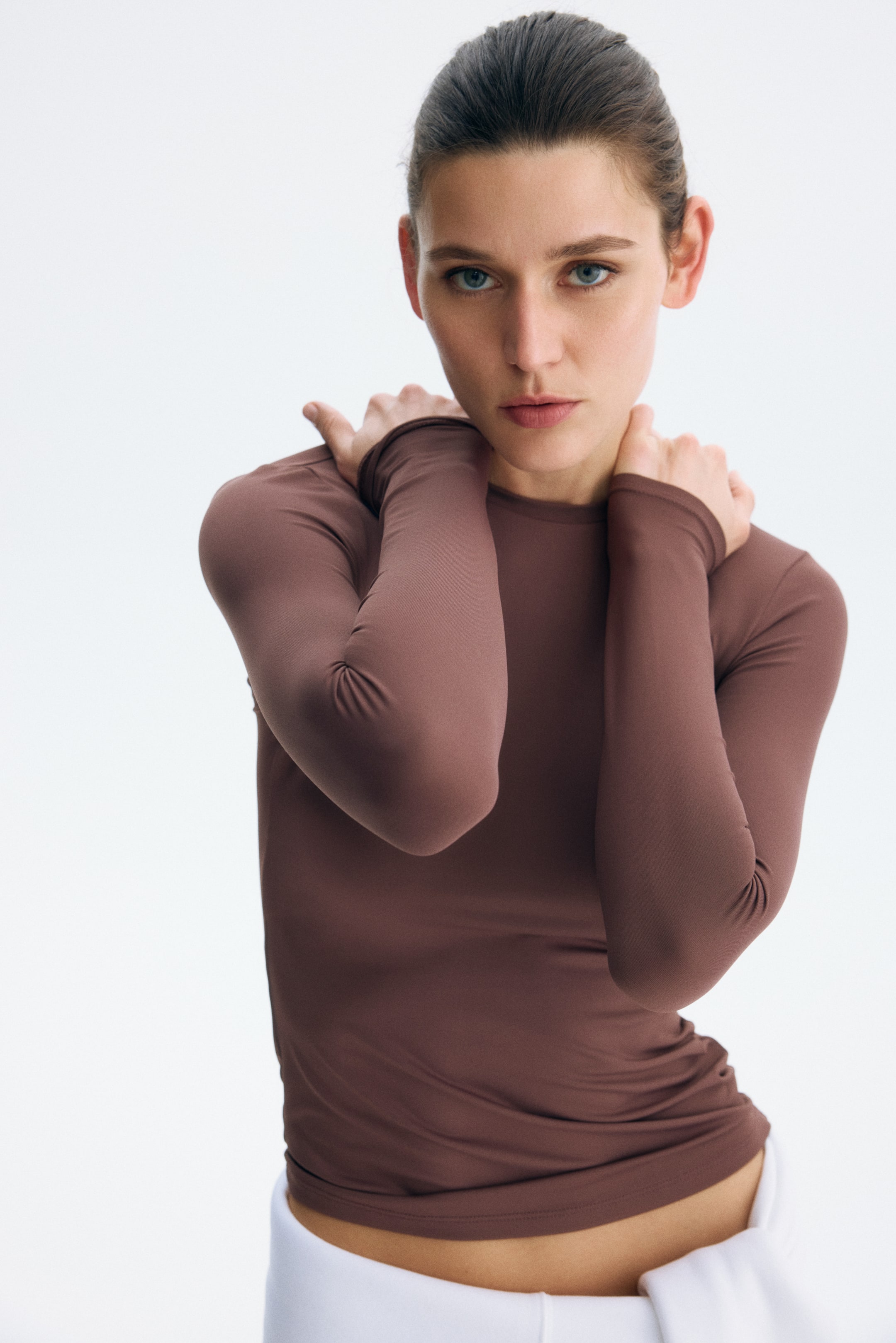 Ingrandisci l'immagine: Una maglia marrone tenue a maniche lunghe aderisce al corpo, vista da un leggero angolo frontale. Presenta uno scollo rotondo e maniche lunghe, con l'orlo che si ferma appena sopra la vita.