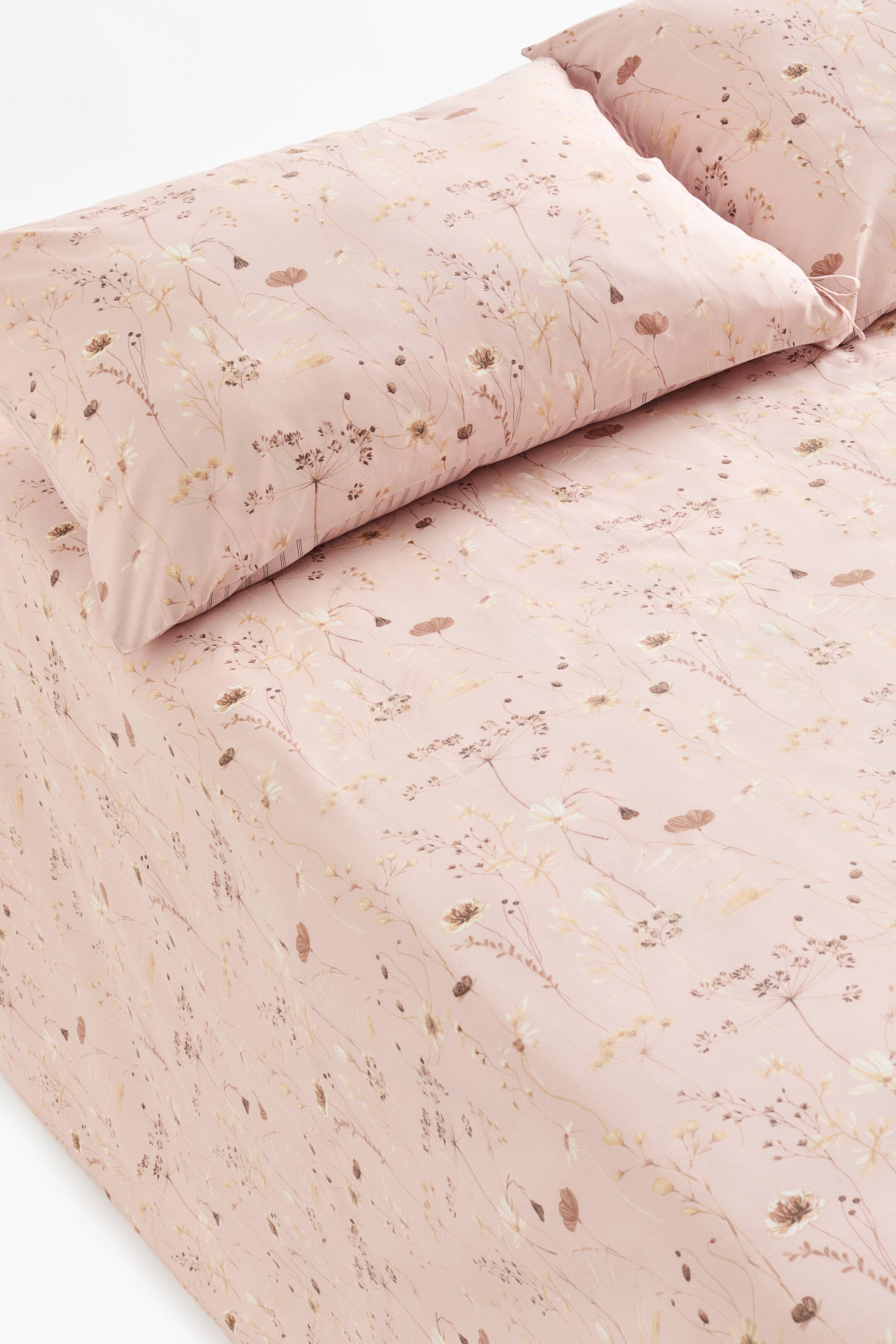 View larger image: A light pink bed sheet set features a flat sheet and two matching pillowcases, all showcasing a delicate floral pattern with small flowers and stems in brown, beige, and white tones. The flat sheet is draped over a bed, angled from the upper left, and one pillowcase shows tie closures on its visible side, facing towards the right.
