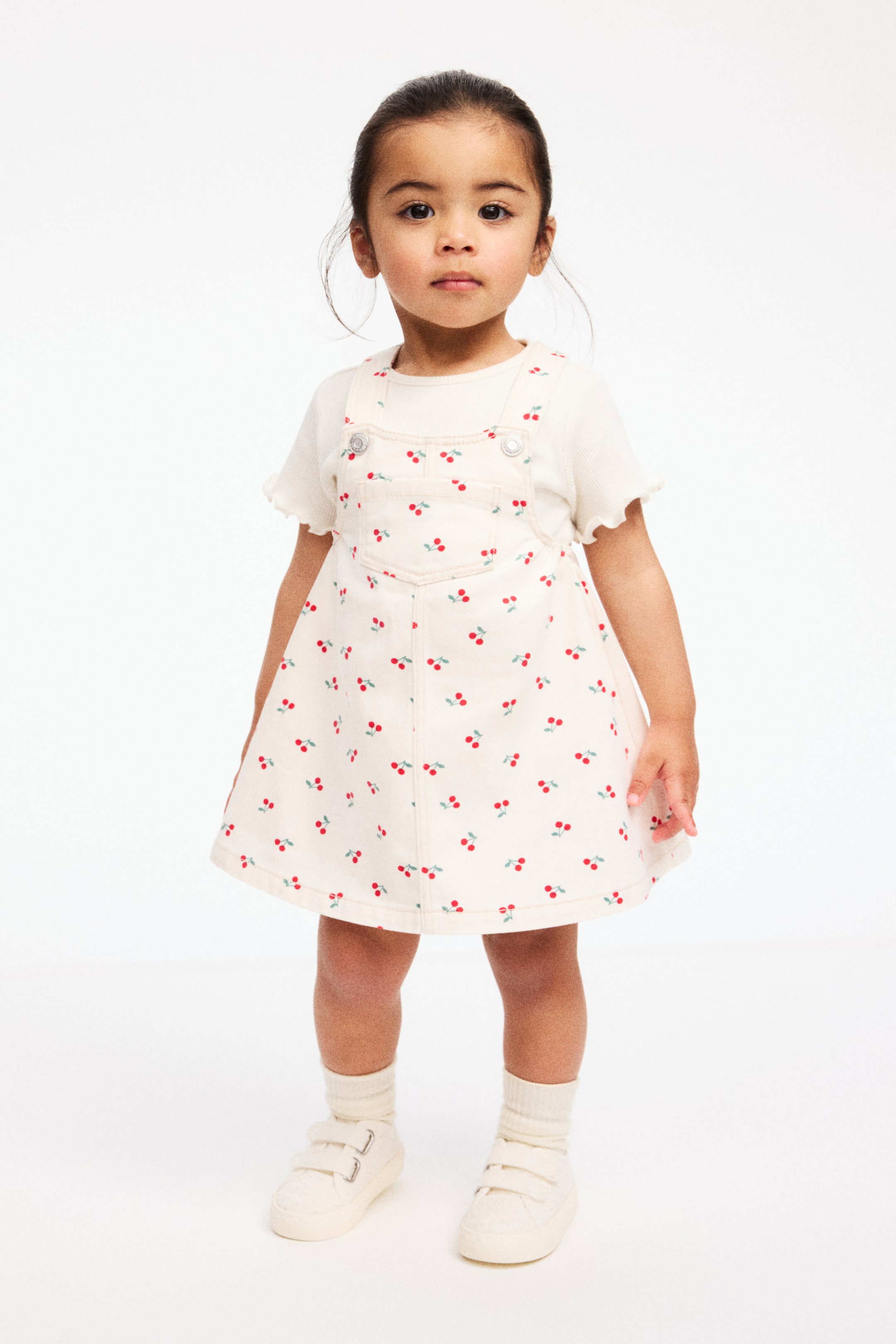 הצגת תמונה גדולה יותר: שמלת אוברול בצבע שמנת‑אוף‑וויט, עם דוגמת דובדבנים אדומים וגבעולים ירוקים, נלבשת מעל טי‑שירט קרם בריב. השמלה כוללת חזית סינר עם כיס טלאי ורצועות כתפיים שנסגרות בעזרת כפתורי מתכת, והטי‑שירט מציג שרוולים קצרים עם וולנים.