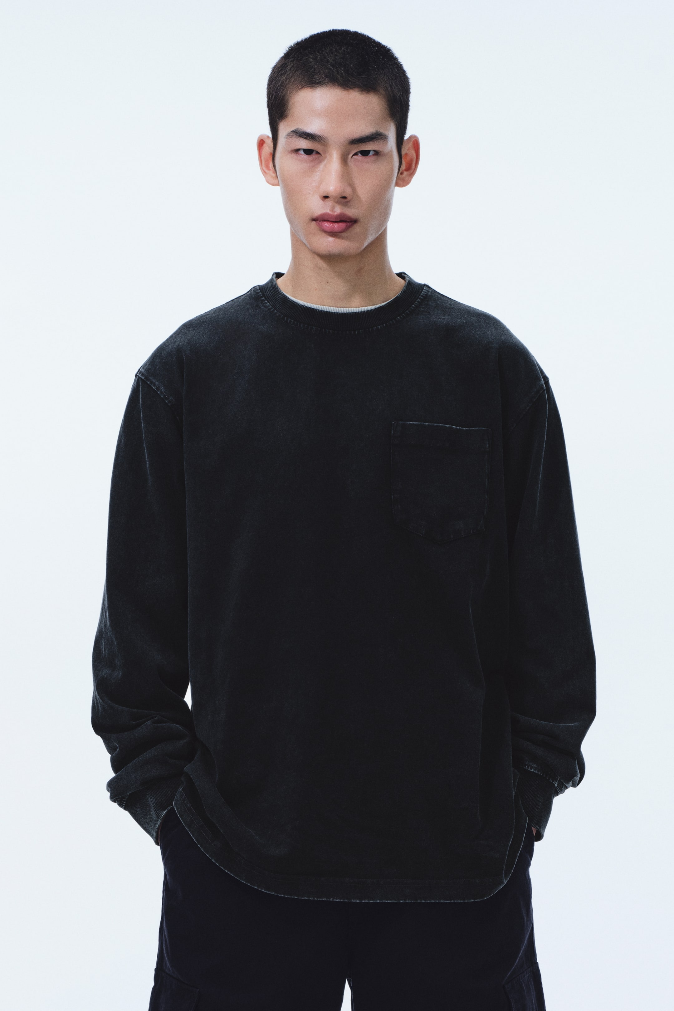 View larger image: A washed black long-sleeved top features a crew neck and a relaxed fit. A patch pocket is placed on the left chest, and a light grey undershirt is visible at the neckline.