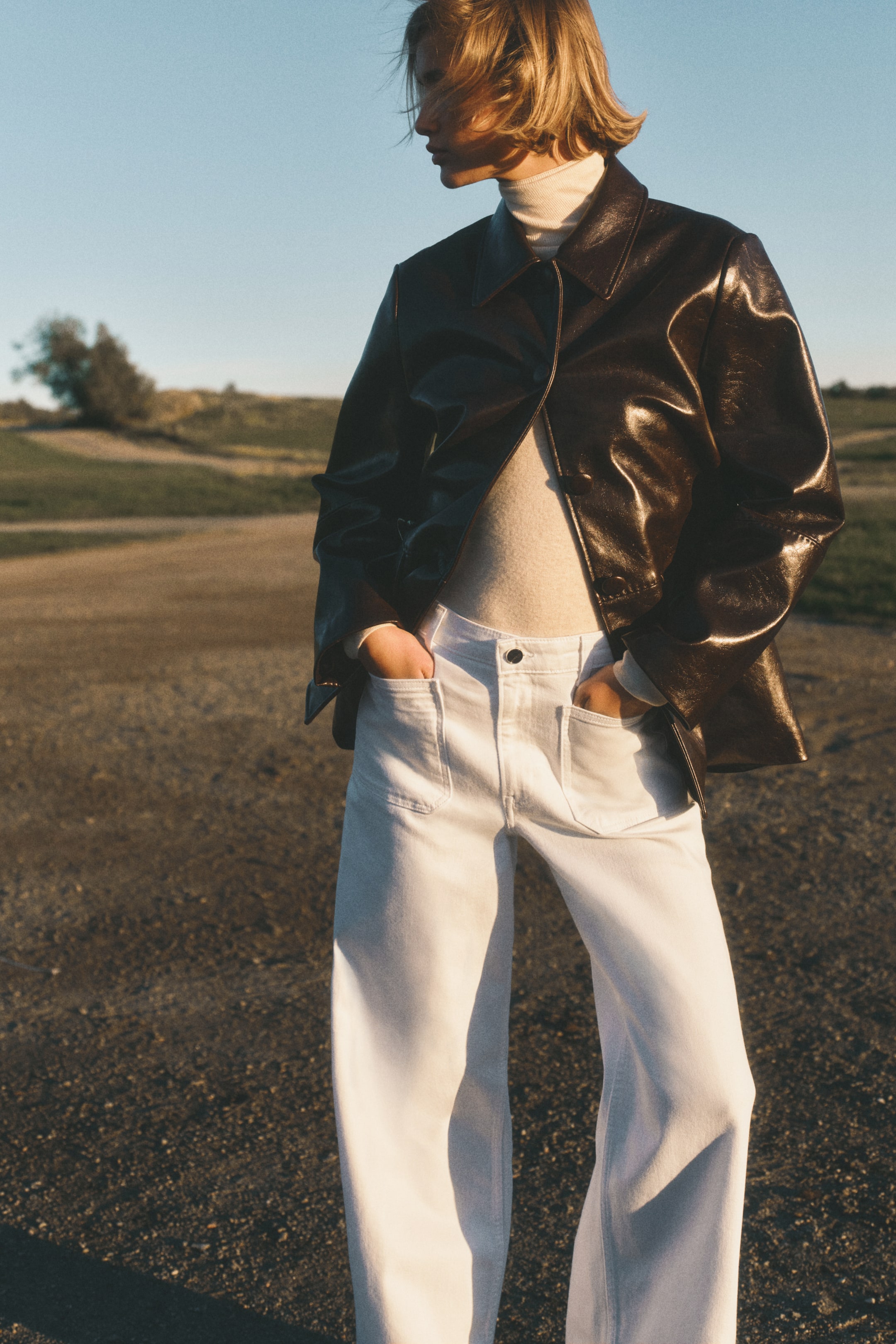 Visualizza immagine più grande: Una giacca marrone scuro e lucida è indossata su un dolcevita a coste color crema chiaro, abbinata a pantaloni a gamba larga color bianco sporco con tasche frontali applicate.