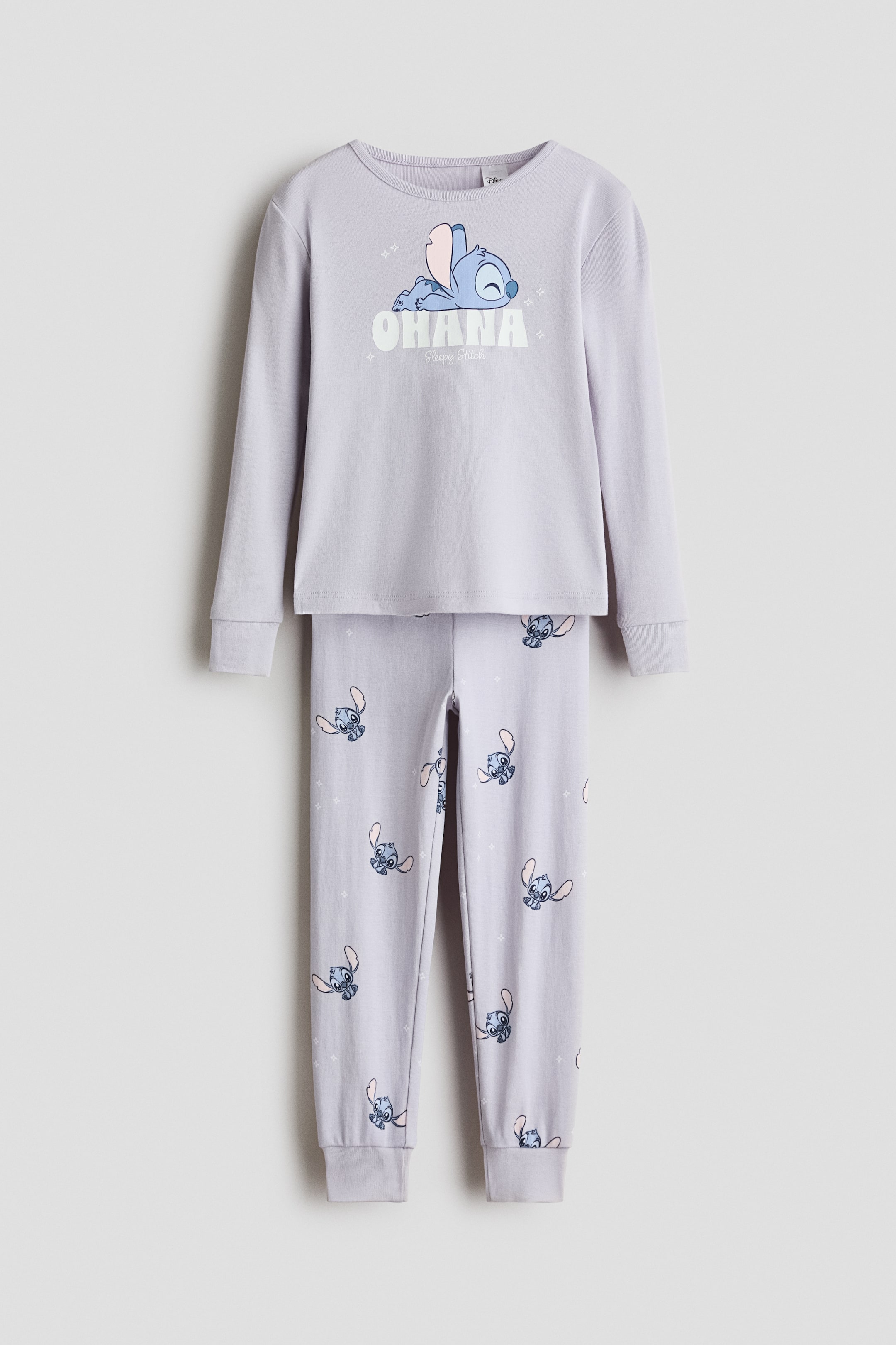 View larger image: A two-piece light purple pyjama set is presented front-facing. The long-sleeved top features a round neck and ribbed cuffs, with a graphic print of a sleeping blue character positioned above the word "OHANA" and "Sleepy Stitch" on the front. The matching long pants showcase an all-over print of the same sleeping blue character, finished with ribbed cuffs.