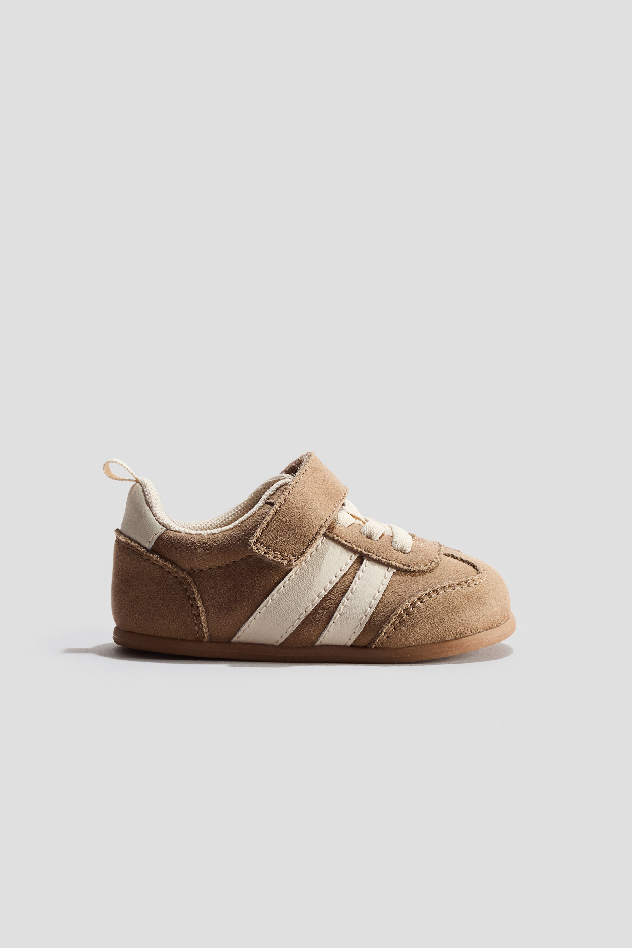 View larger image: Trainers. This tan suede trainer is showcasing its side profile, highlighting two off-white stripes, an off-white heel pull tab, and a single hook-and-loop strap fastening over off-white faux laces. It is positioned on a flat brown sole.