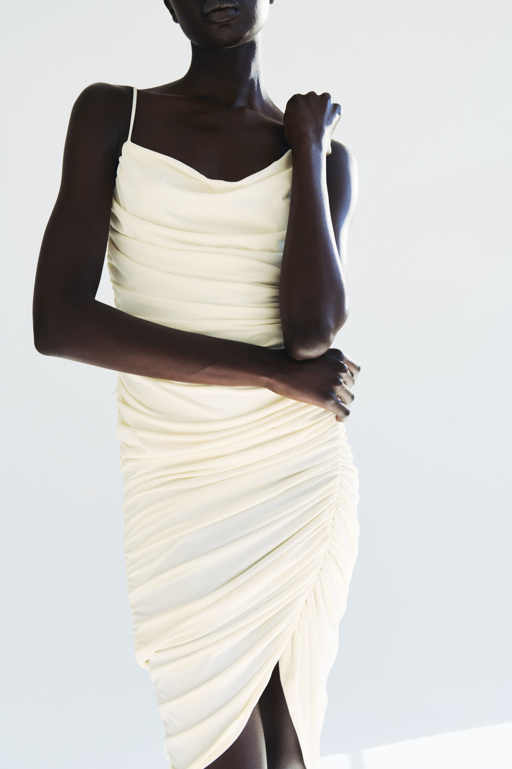 View larger image: The model is wearing a cream, knee-length fitted dress featuring a cowl neckline and spaghetti straps. The dress showcases gathered seams that create a draped effect, along with a slit on one side of the front. The model has their arms crossed, with one hand resting on the upper arm and the other on the hip.