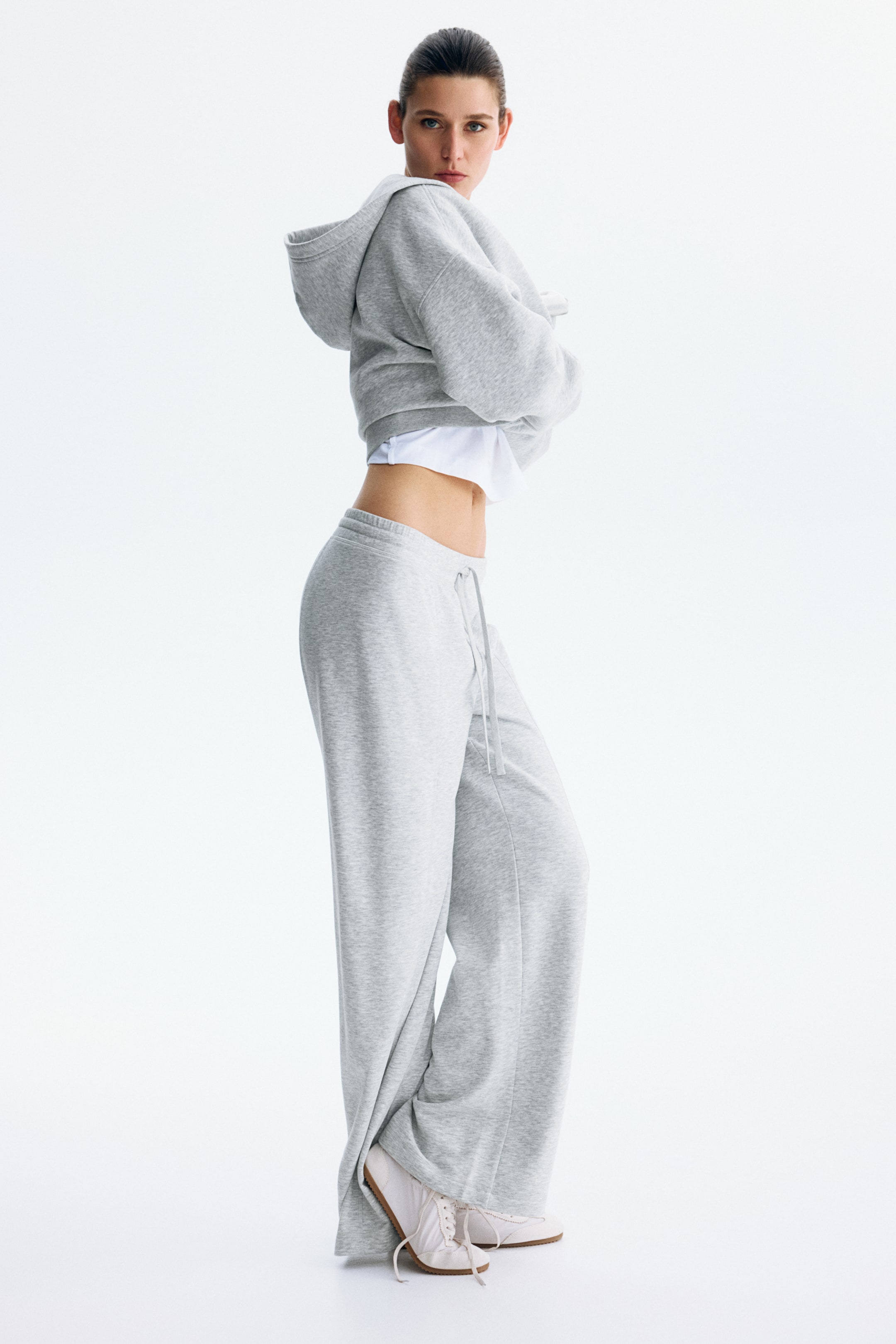 View larger image: The gray cropped hoodie layers over a white top, featuring long sleeves and a hood. The matching gray wide-leg sweatpants display a visible drawstring at the waist. This athletic set is shown from a three-quarter side angle, highlighting its relaxed fit.