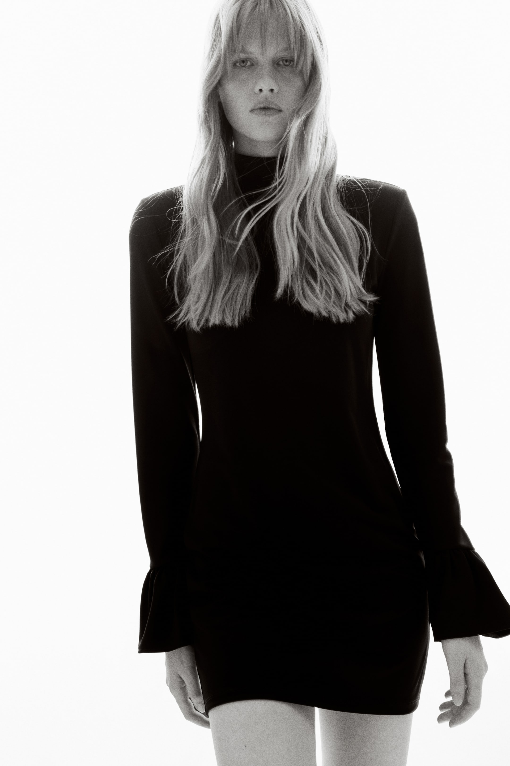 Zobrazit větší obrázek: Krátké černé šaty na tělo, které mají rolák, dlouhé rukávy s širokým volánem na manžetách a výraznou linii ramen, nosí modelka. Modelka stojí a předvádí šaty.