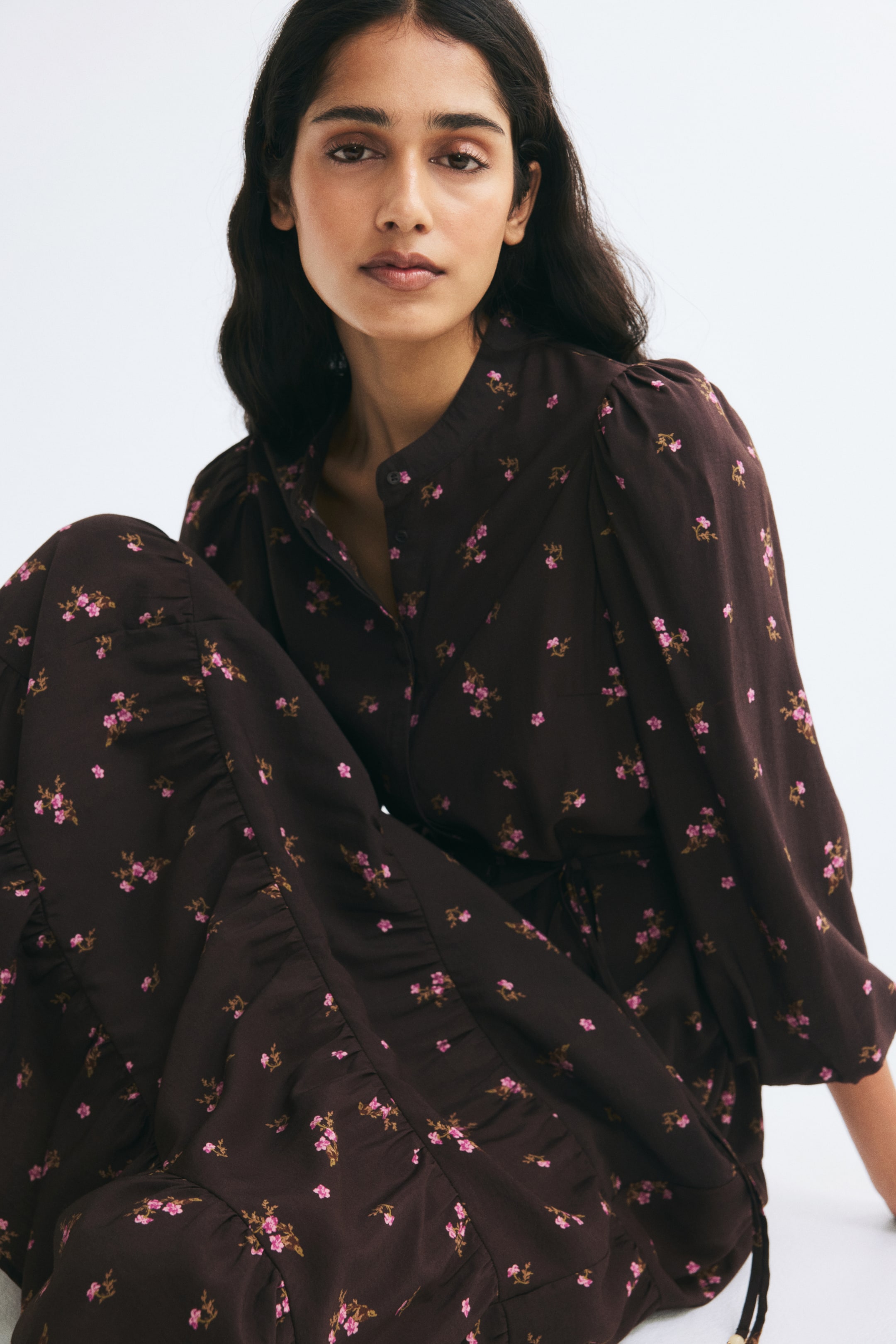 View larger image: A long, relaxed-fit dress in a dark brown floral print is shown from the front, featuring a band collar with a partially open button placket. The dress displays three-quarter-length puff sleeves with pleated cuffs and a flared skirt with gathered tiers.