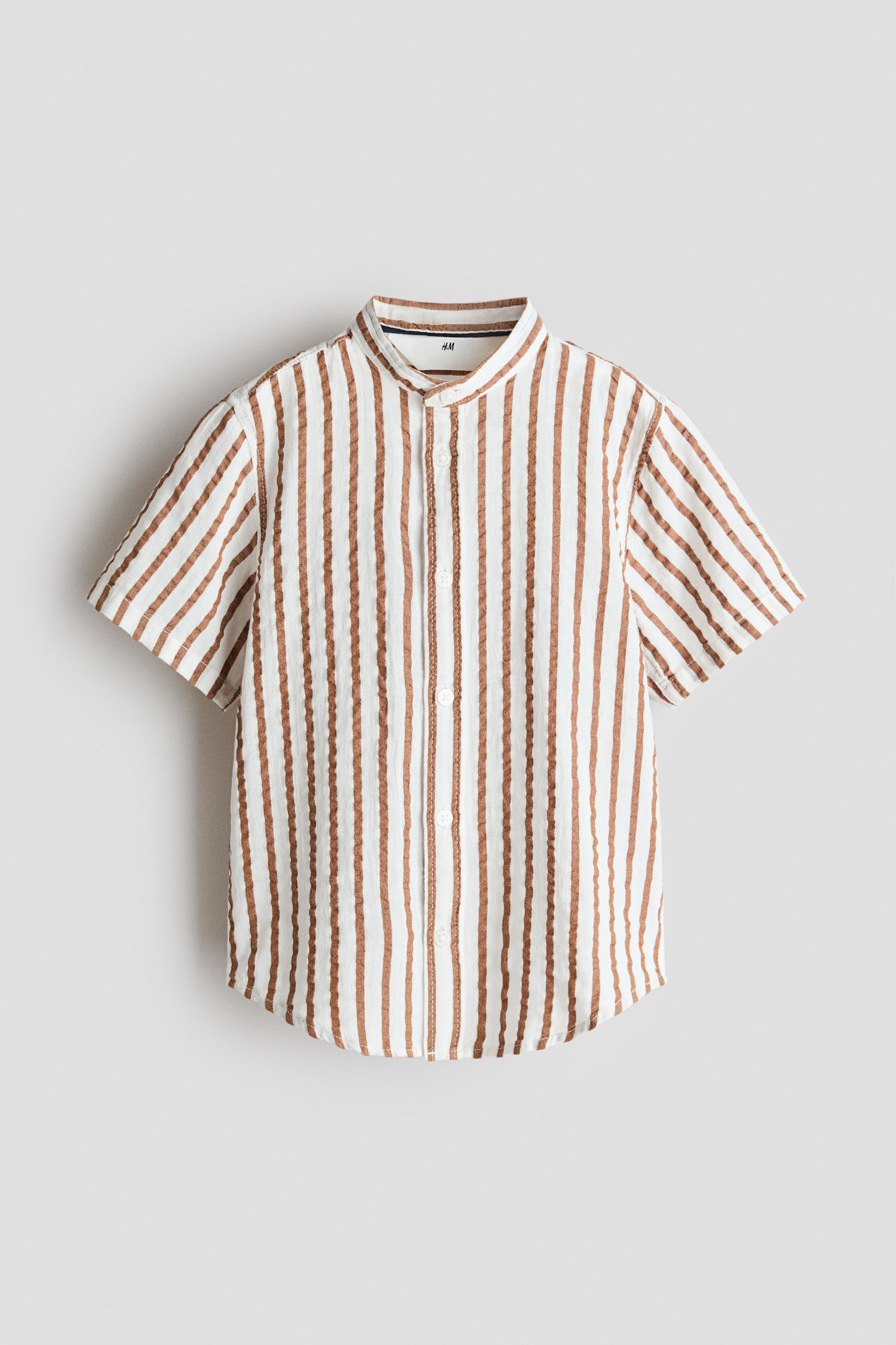 View larger image: Short-sleeved shirt displayed frontally, showcasing white fabric adorned with textured brown vertical stripes. It boasts a band collar, a full-length button placket with white buttons, and a rounded hem.