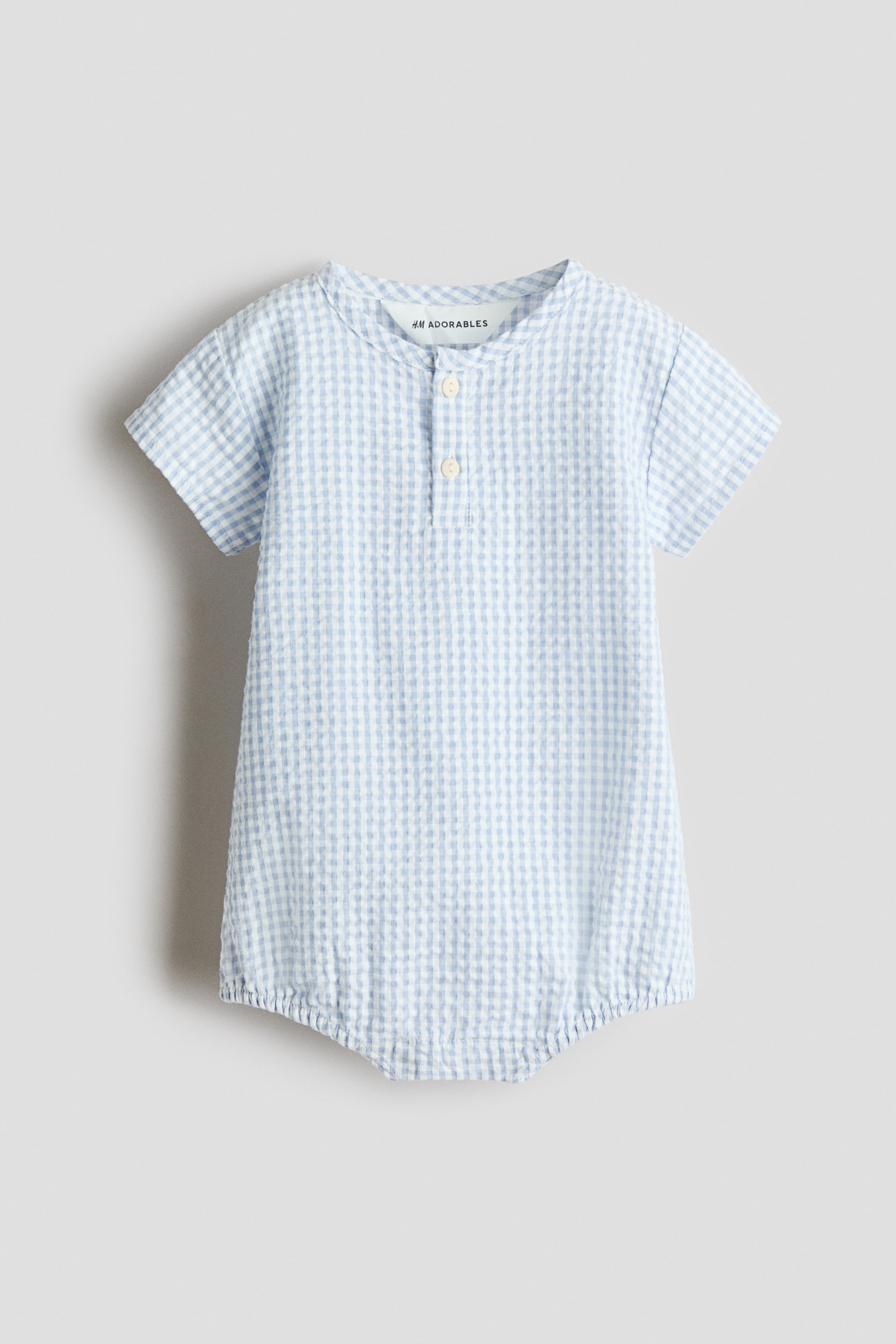 View larger image: A front-facing baby romper showcasing a light blue and white gingham pattern. It is crafted with a band collar, a two-button henley placket, short sleeves, and elasticated leg openings, giving it a bubble shape.