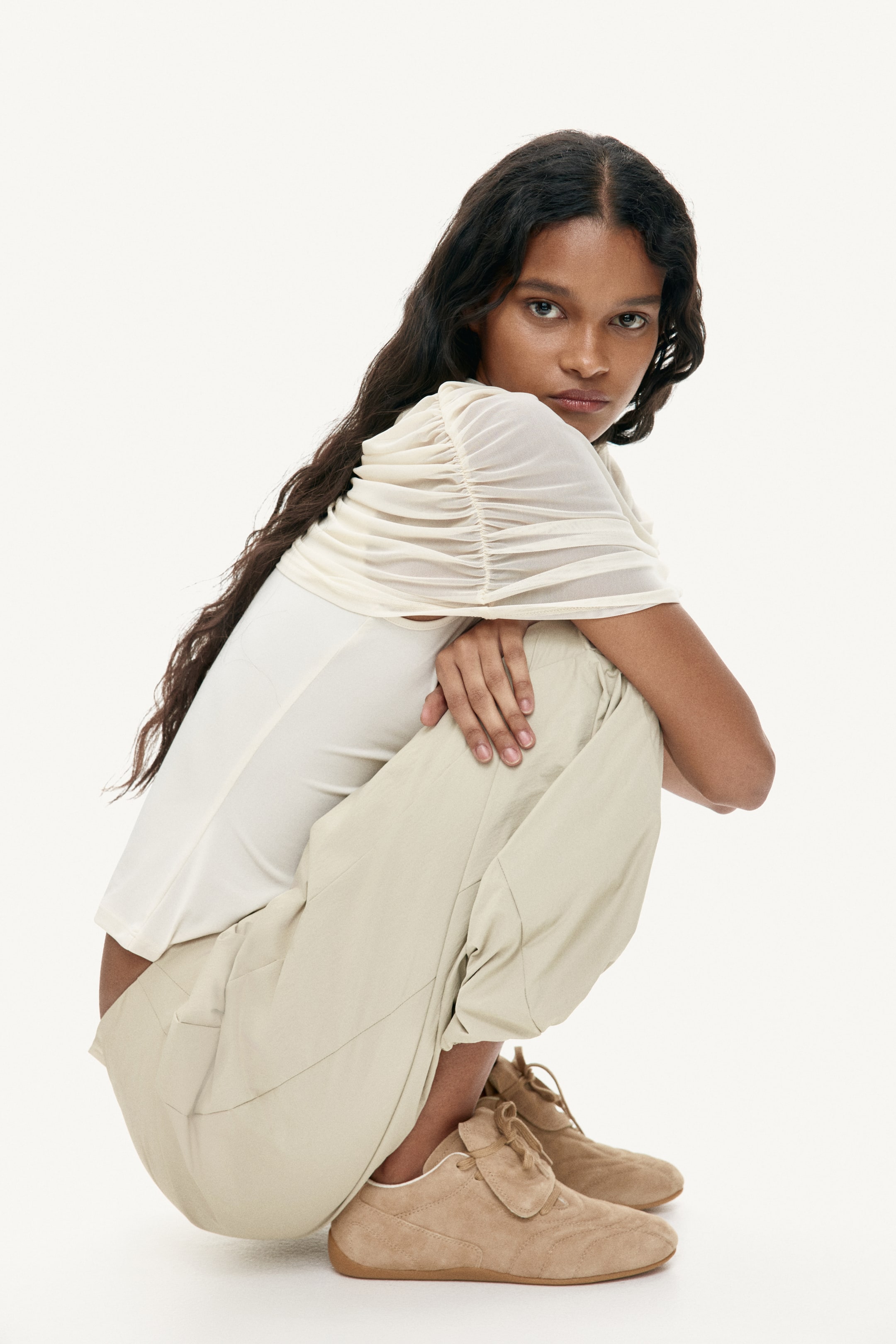 Grösseres Bild ansehen: Ein cremefarbenes Mesh-Top mit umfangreichen Raffungen, die einen gerafften Effekt über die Schultern und Oberarme hinweg erzeugen. Es ist über einem festen cremefarbenen Kleidungsstück geschichtet, das am Oberkörper darunter sichtbar ist.