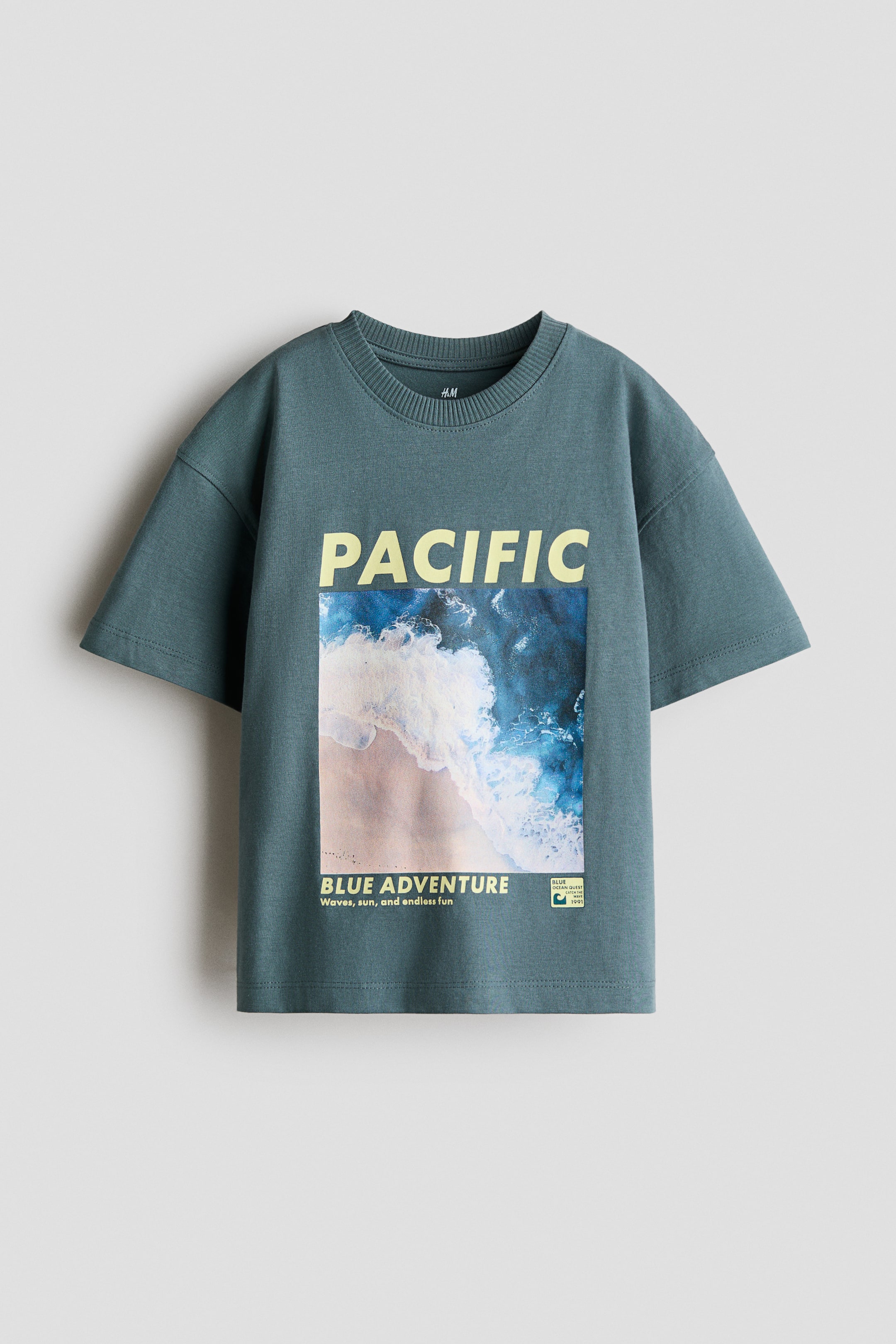 Ver imagen más grande: Una camiseta de corte holgado se presenta de frente, con un color verde azulado oscuro, un cuello redondo con ribete y hombros caídos. Un destacado estampado gráfico rectangular adorna el frente, mostrando una vista aérea de olas azules del océano sobre una playa de arena, con la palabra "PACIFIC" arriba y "BLUE ADVENTURE" abajo.