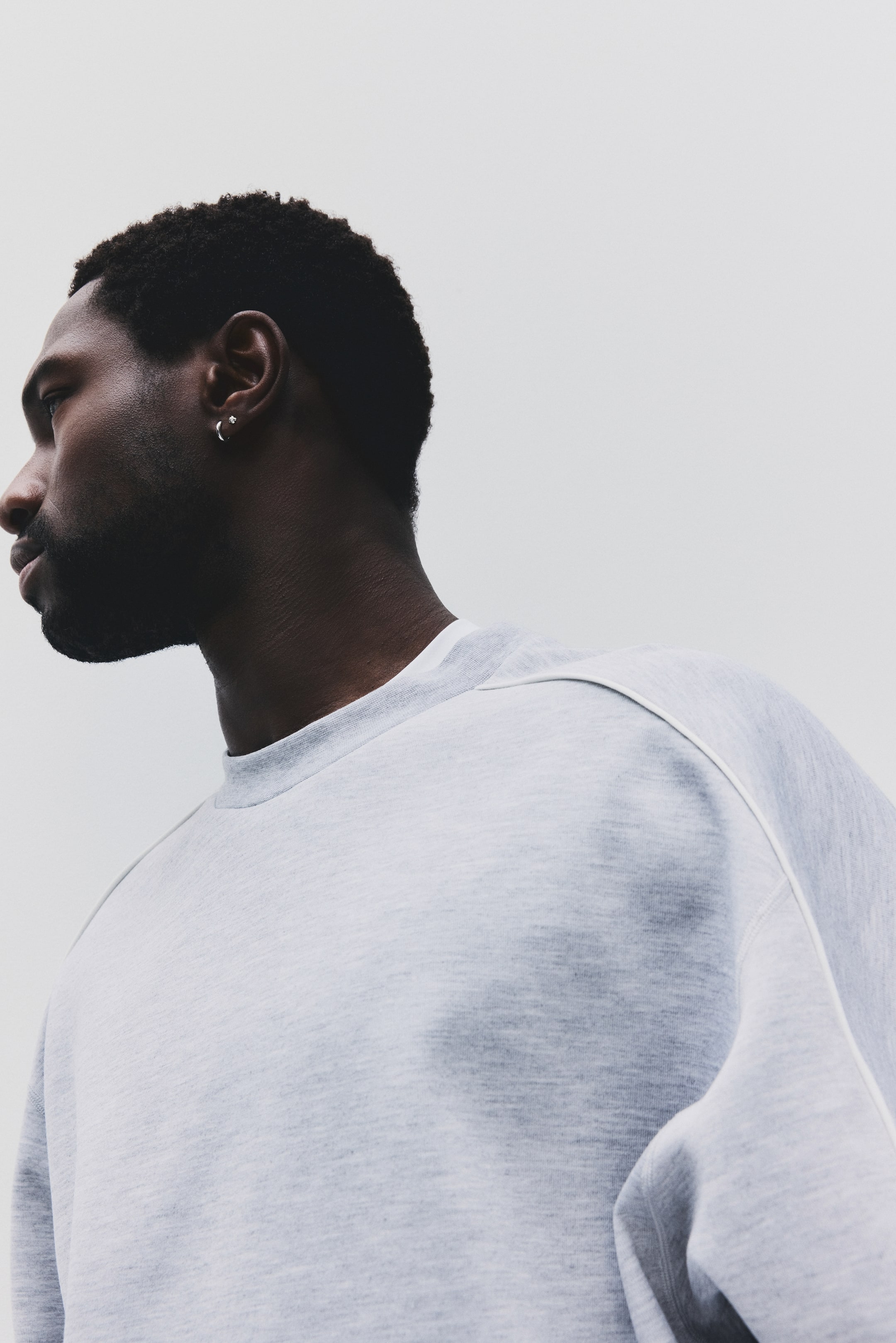 Visa större bild: En ljusgrå sweatshirt med rund hals bärs, vinklad åt vänster, som visar en framträdande design-söm som löper från kragen över vänster axel och ner längs ärmen. Sweatshirten har en avslappnad passform och är tillverkad i ett melerat grått tyg.