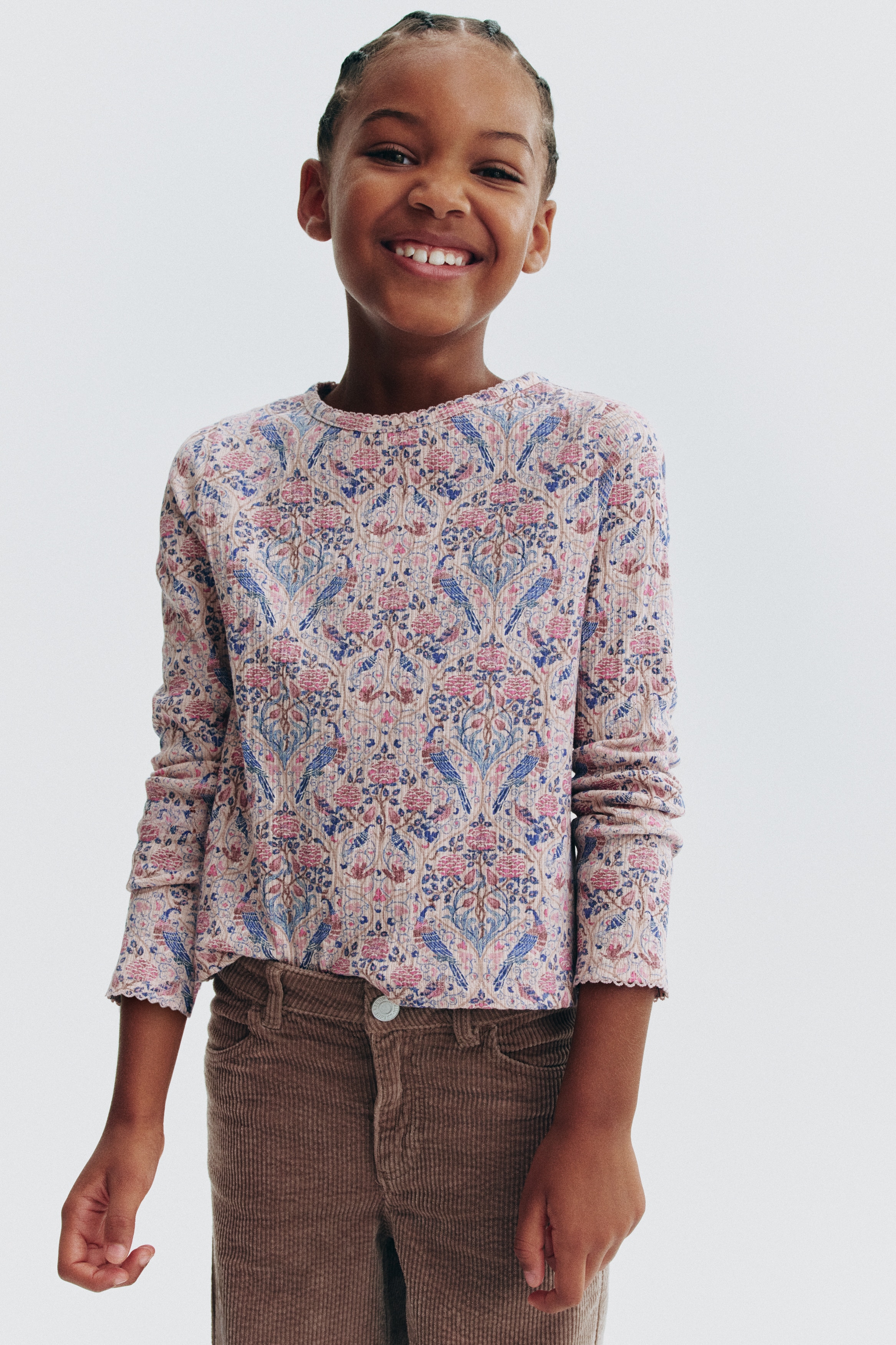 A long-sleeve top made from pink ribbed cotton jersey is shown from the front, featuring an all-over floral and bird print in shades of pink and blue. The top displays a round neckline and cuffs, both detailed with a delicate picot trim.