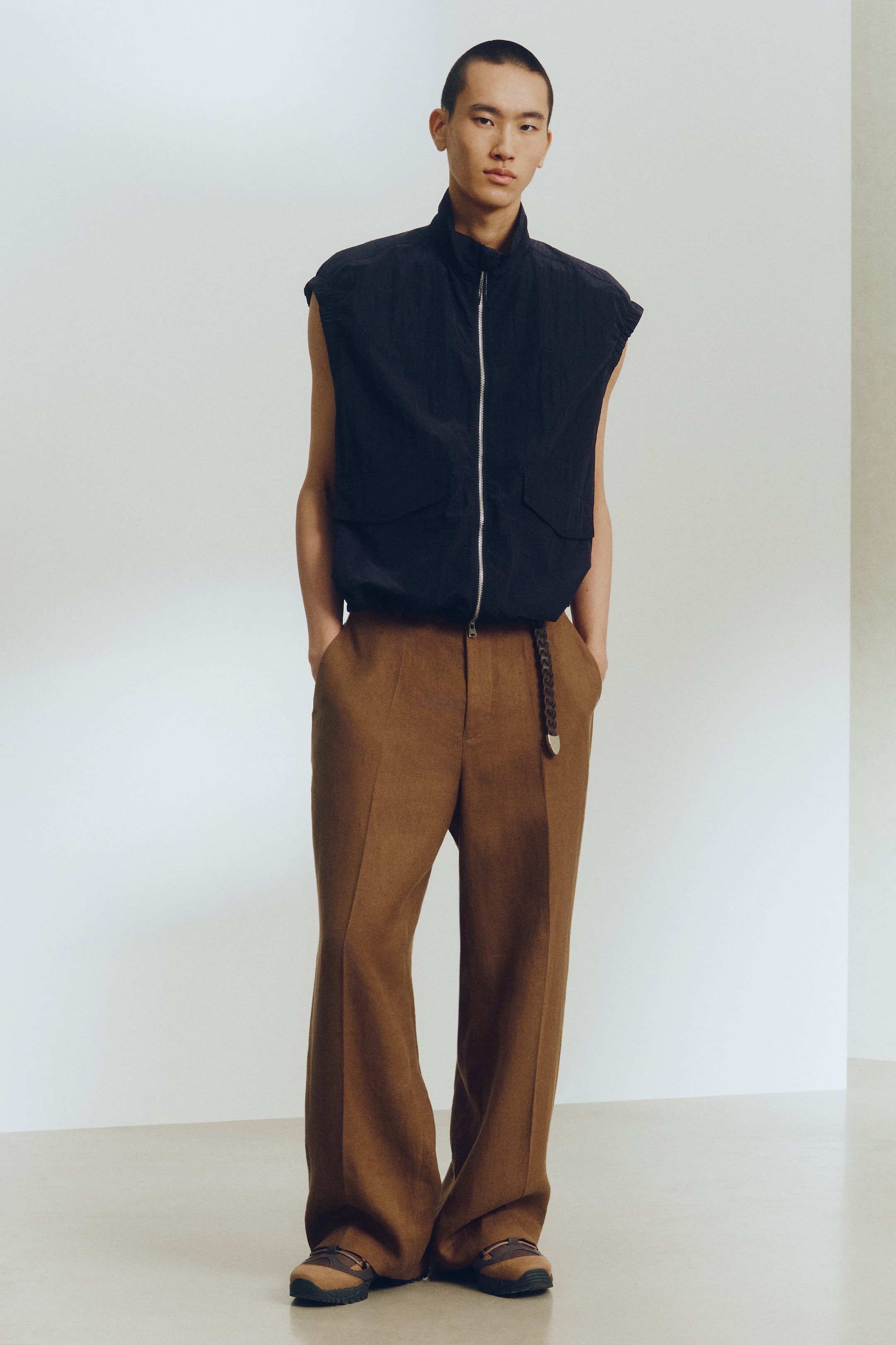 Zobacz większy obraz: Czarna, marszczona kamizelka bez rękawów z kołnierzem stojącym, przednim zamkiem błyskawicznym i dwoma kieszeniami z klapkami na piersi, noszona jest na brązowych spodniach z szerokimi nogawkami. Spodnie są stylizowane z brązowym plecionym paskiem i lekko opadają na brązowe i beżowe buty wsuwane.