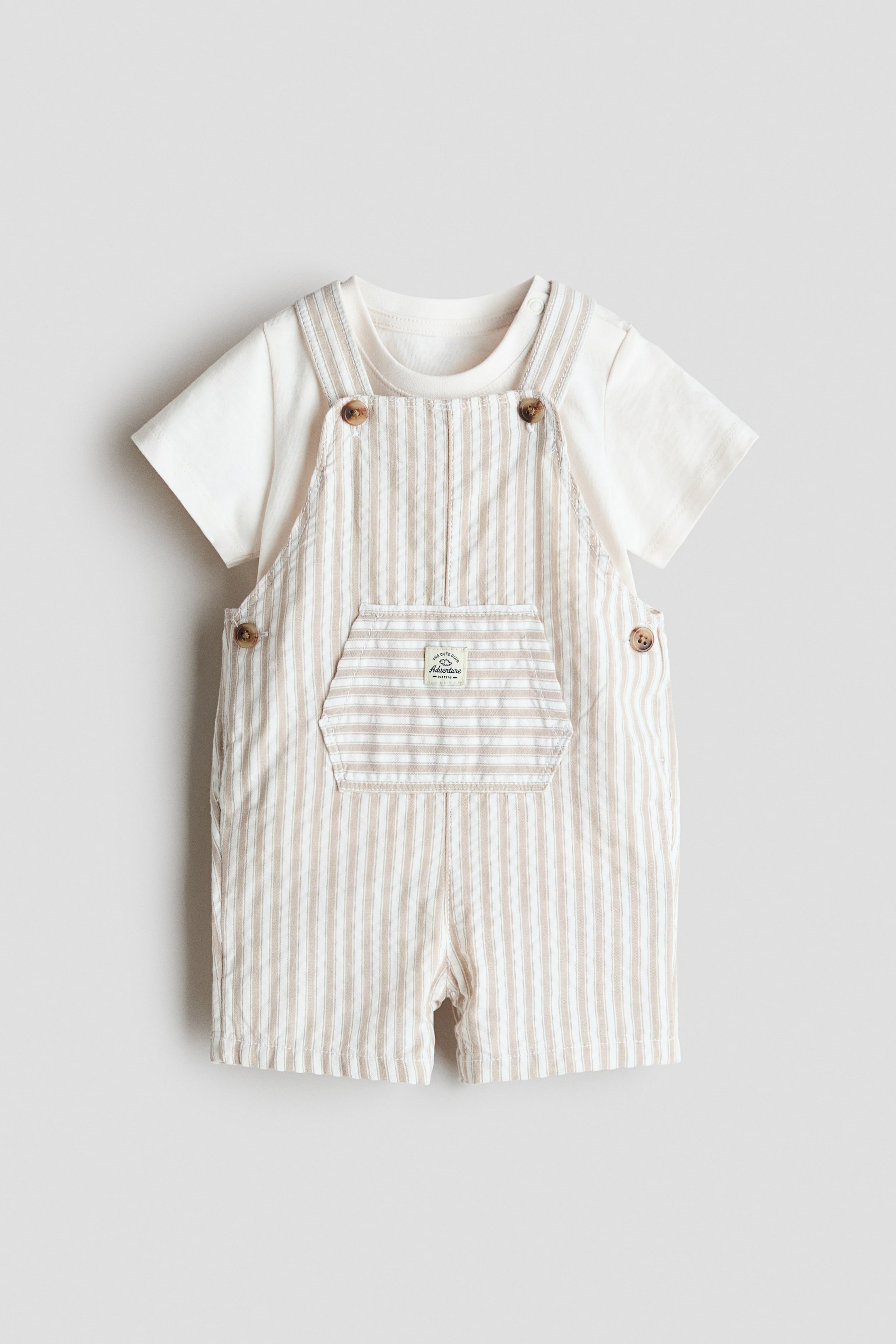 Agrandir l'image: Un t-shirt à manches courtes pour bébé, de couleur blanc cassé, est superposé sous une salopette courte rayée, tous deux orientés vers l'avant. La salopette présente des rayures verticales texturées blanches et beiges, des bretelles réglables fixées par des boutons marron, et une poche poitrine hexagonale.