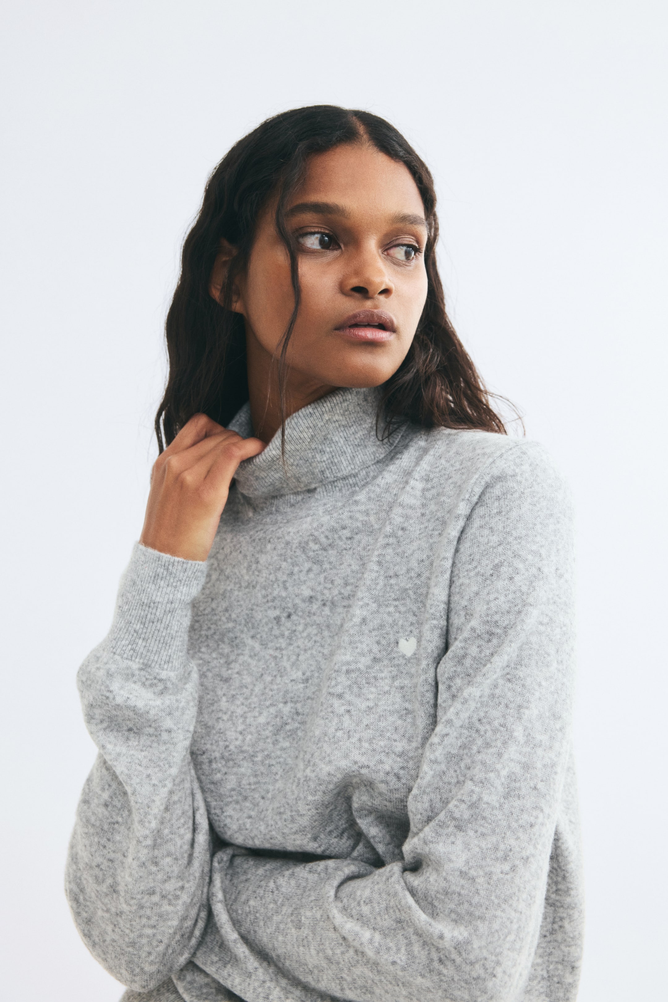 View larger image: A light grey, soft knit sweater featuring a ribbed turtleneck and long sleeves with ribbed cuffs is being worn by the model. The model is adjusting the collar of the sweater, which also features a small, light-coloured heart detail on the left chest.