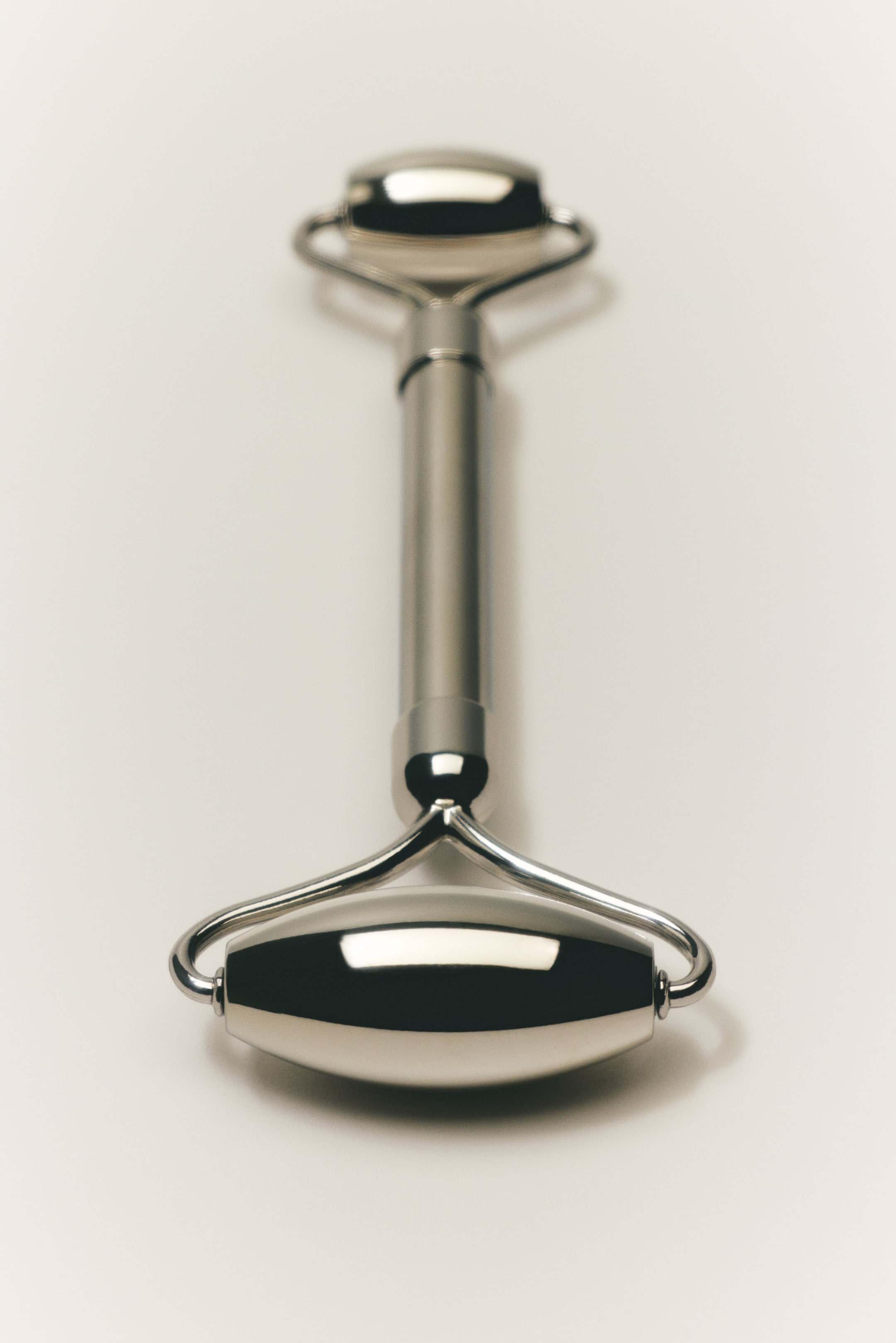 Ampliar la imagen: Un rodillo de belleza metálico yace en posición horizontal, con dos rodillos pulidos de forma ovalada, cada uno alojado en un armazón curvo de metal plateado que se conecta al mango cilíndrico central.