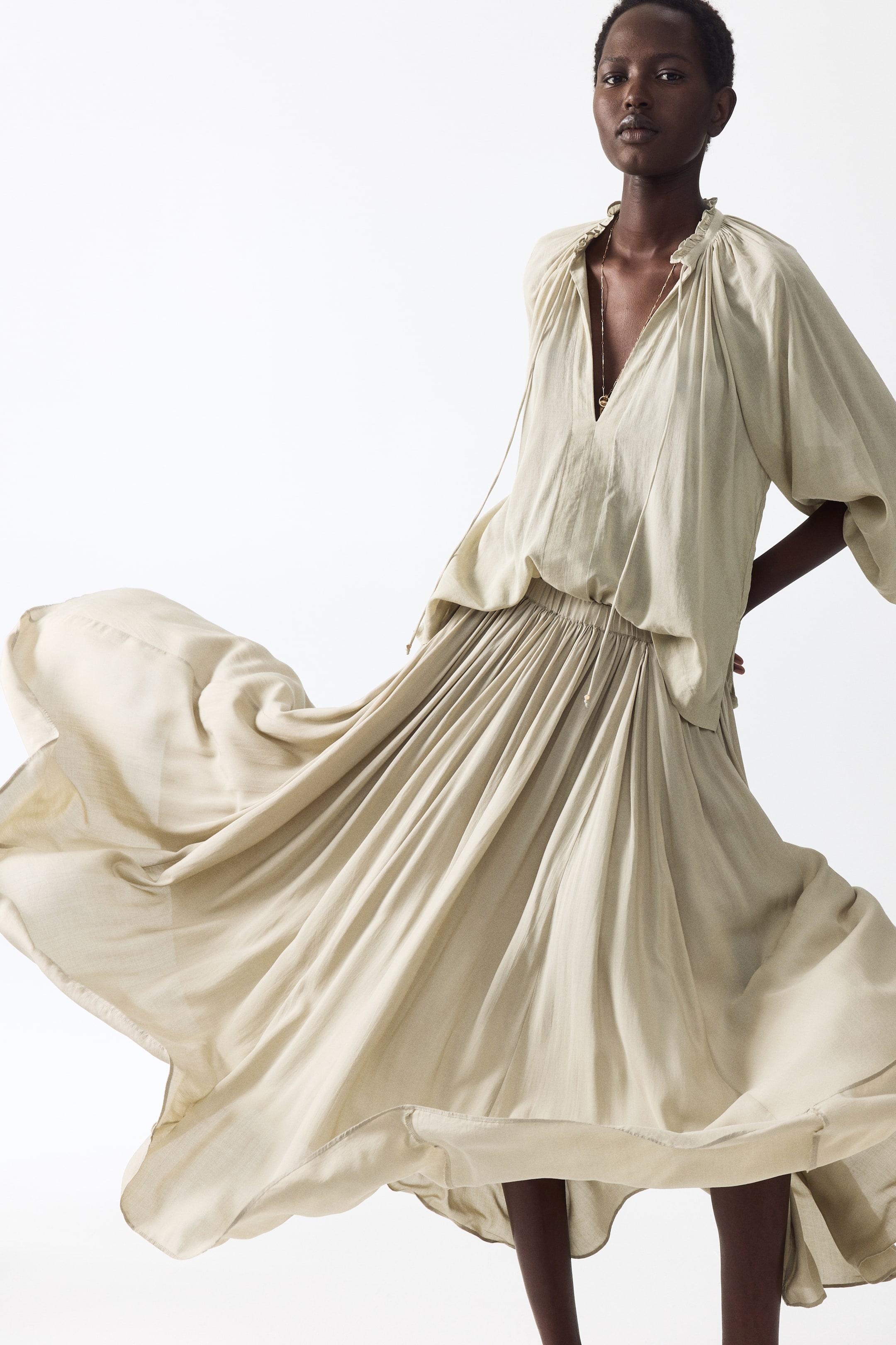 View larger image: The light beige flowing blouse features a ruffled V-neck with drawstrings and wide, voluminous sleeves. Its hem is tucked into a matching maxi skirt, which showcases a gathered waist and a full, flowing hem, creating a voluminous silhouette.