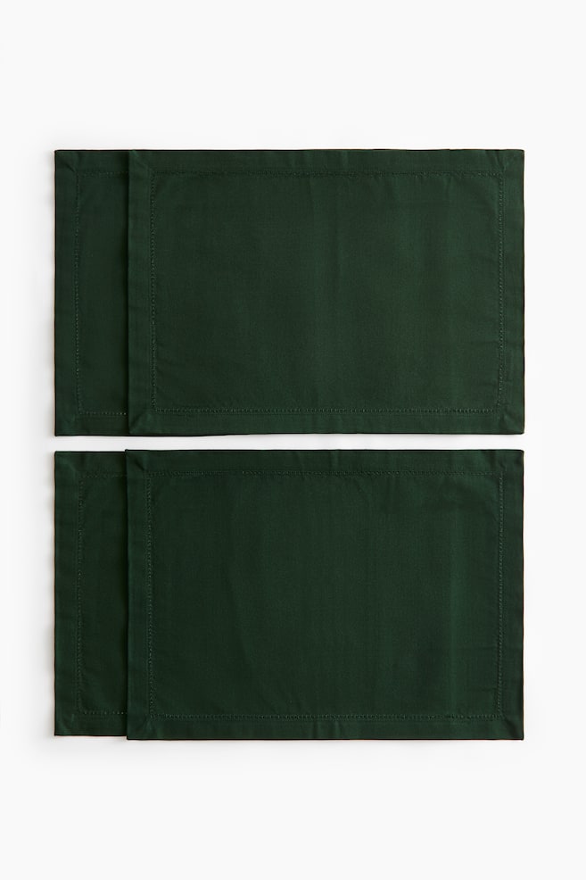 Quatre sets de table en toile de coton vert foncÃ© sont posÃ©s Ã  plat, vus de face, disposÃ©s en deux paires lÃ©gÃ¨rement dÃ©calÃ©es afin de mettre en valeur leurs larges bordures et les dÃ©tails de point ajourÃ© le long des bords intÃ©rieurs. - 1