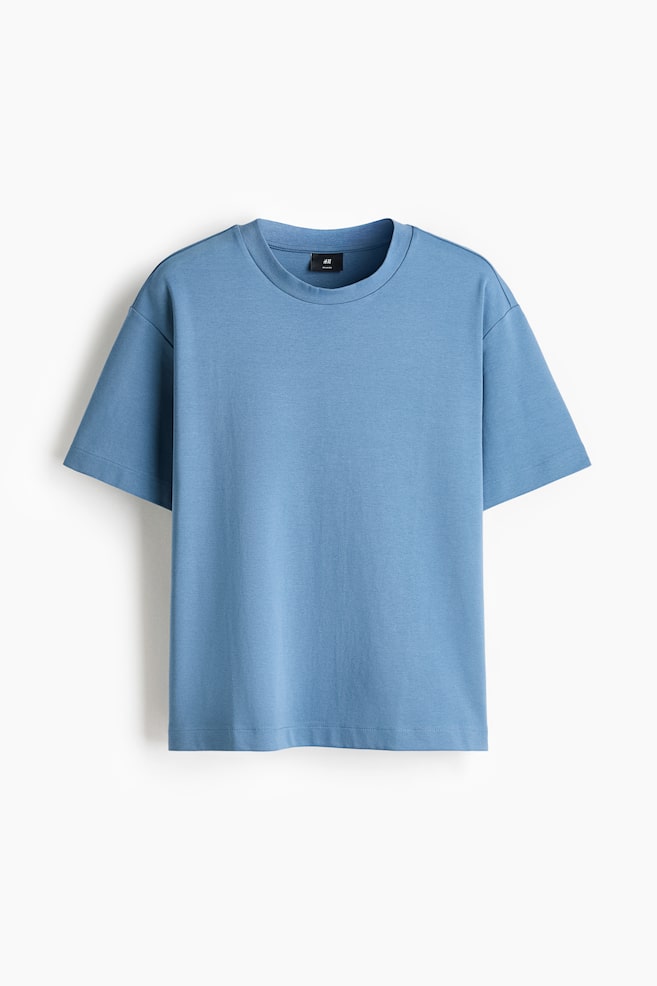 Een lichtblauw T-shirt in boxy-stijl is van voren te zien, met een ronde hals met ribboord, verlaagde schouders en wijde korte mouwen. Het losvallende topje heeft een recht afgewerkte zoom. - 1