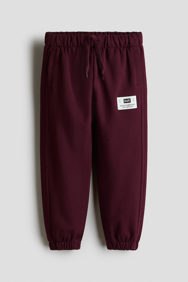 Burgundy track pants are shown from the front, featuring an elasticated waistband with a matching drawstring and side slip pockets. The pants are tapering down to elasticated hems and display a small, white rectangular patch on the upper left thigh. - 1