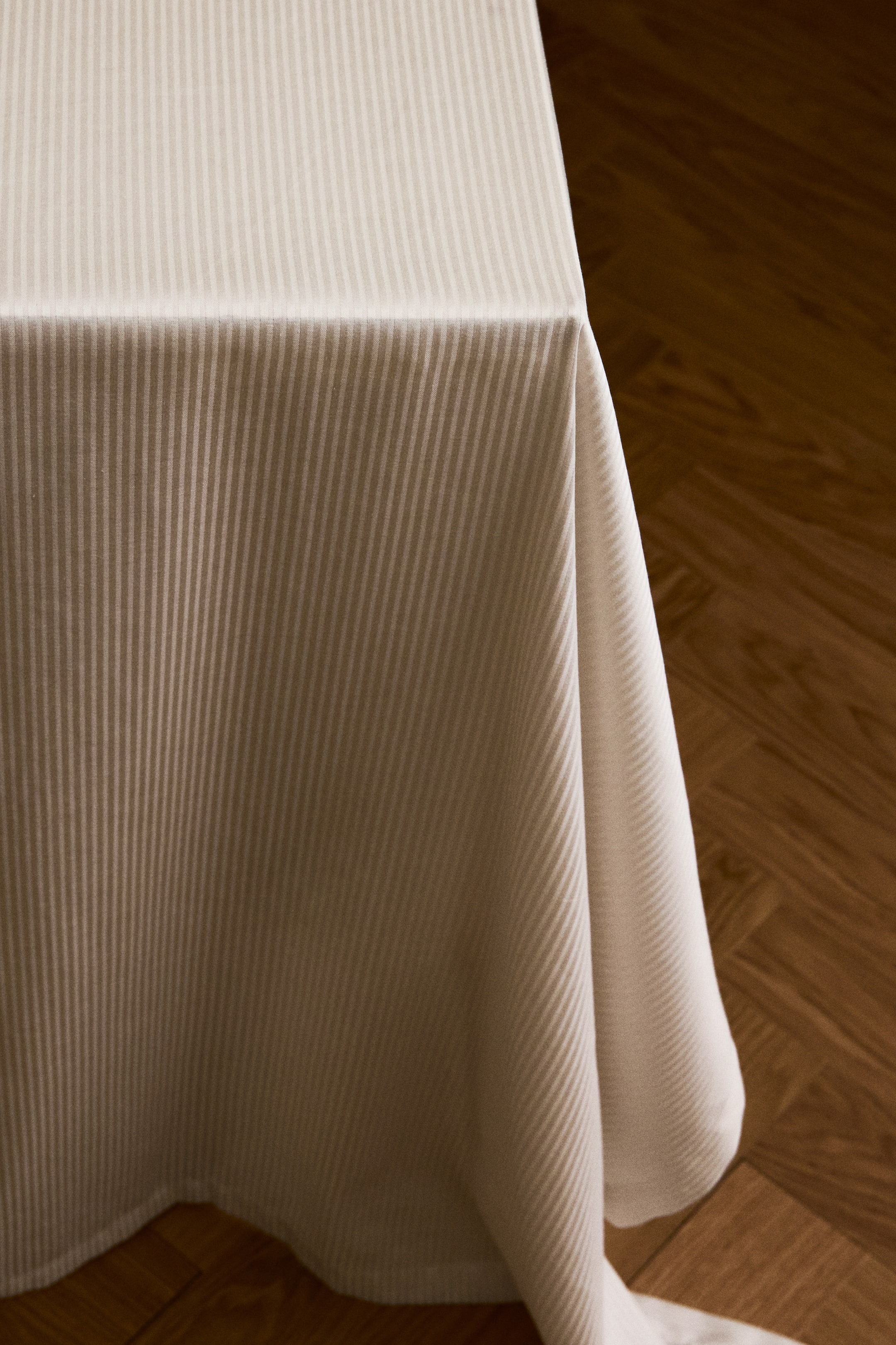 View larger image: A light beige cotton-blend tablecloth featuring a matching-tone, fine-striped woven pattern is draped over a table corner. The fabric falls in structured folds, displaying its smooth texture and finished folded hem.