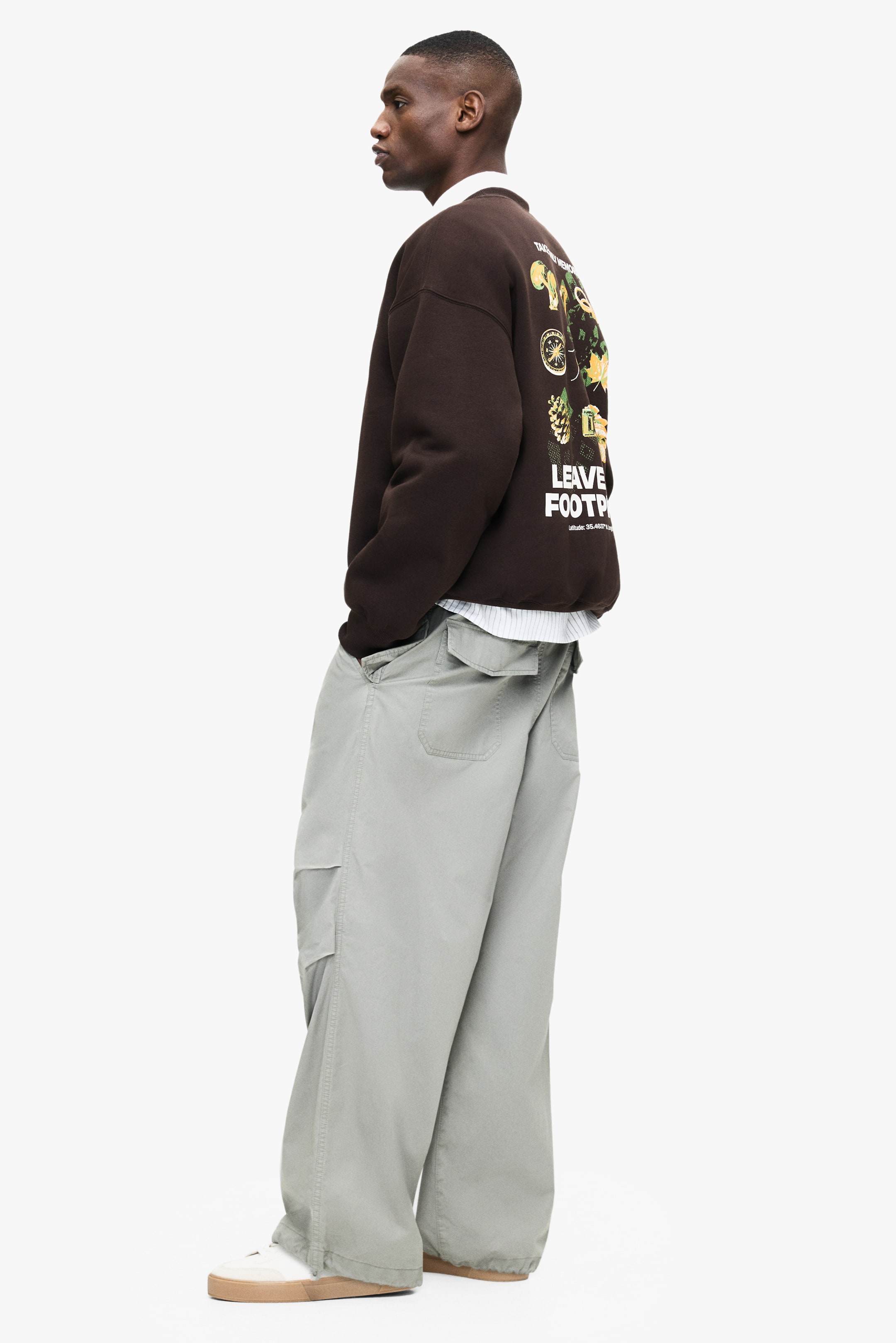 Grösseres Bild ansehen: Ein braunes Rundhals-Sweatshirt mit einem großen Grafikdesign auf dem Rücken, das Text und verschiedene Illustrationen zeigt, wird vom Model getragen und ist über einem weißen Hemd mit Kragen geschichtet.