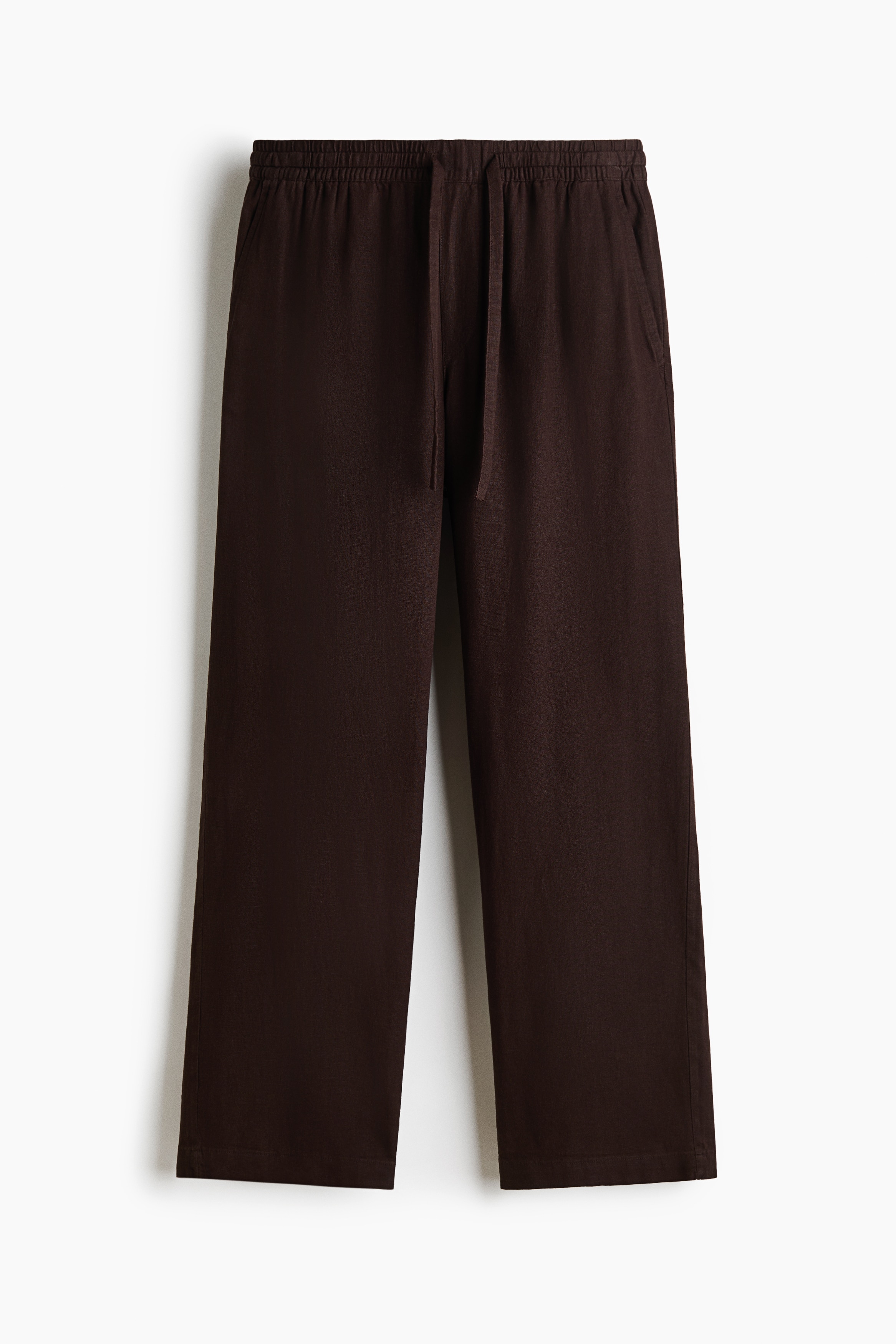 Dark brown, relaxed-fit trousers featuring an elasticated drawstring waist and diagonal side pockets are worn by the model, who is standing with one hand in a pocket. The trousers are styled with a tucked-in black V-neck T-shirt, black shoes, and a brown and tan bag carried under one arm.