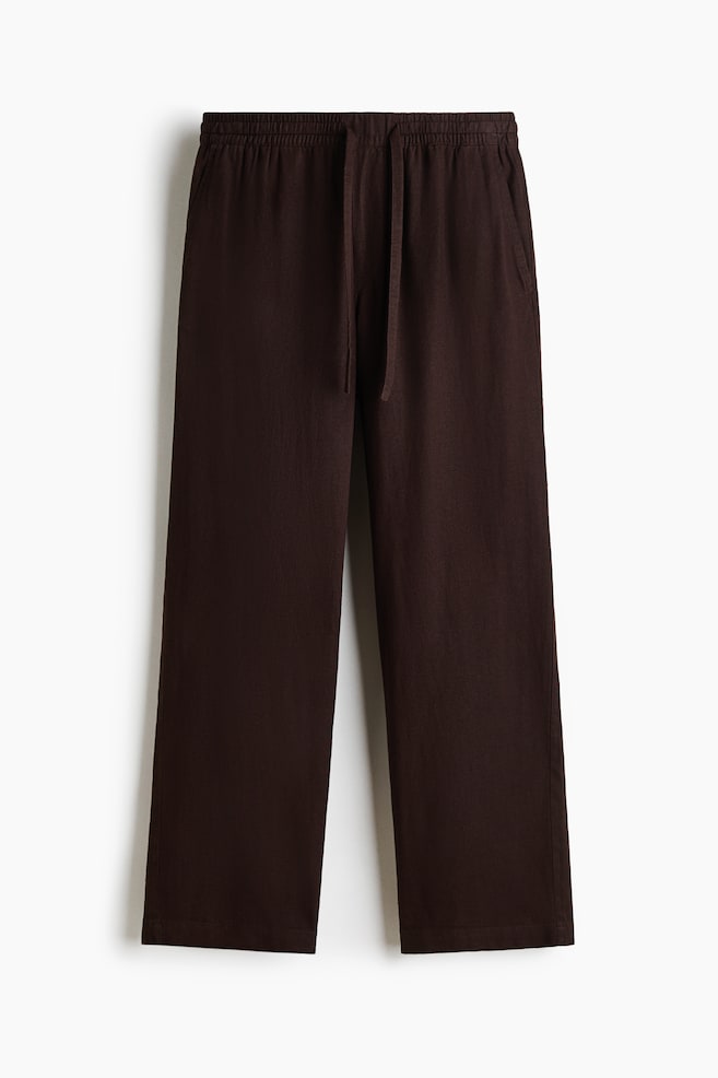 A pair of dark brown, straight-leg trousers is hanging facing the front, showcasing an elasticated waistband with a matching fabric drawstring. The trousers are displaying side slip pockets and a relaxed silhouette crafted from a textured fabric. - 1