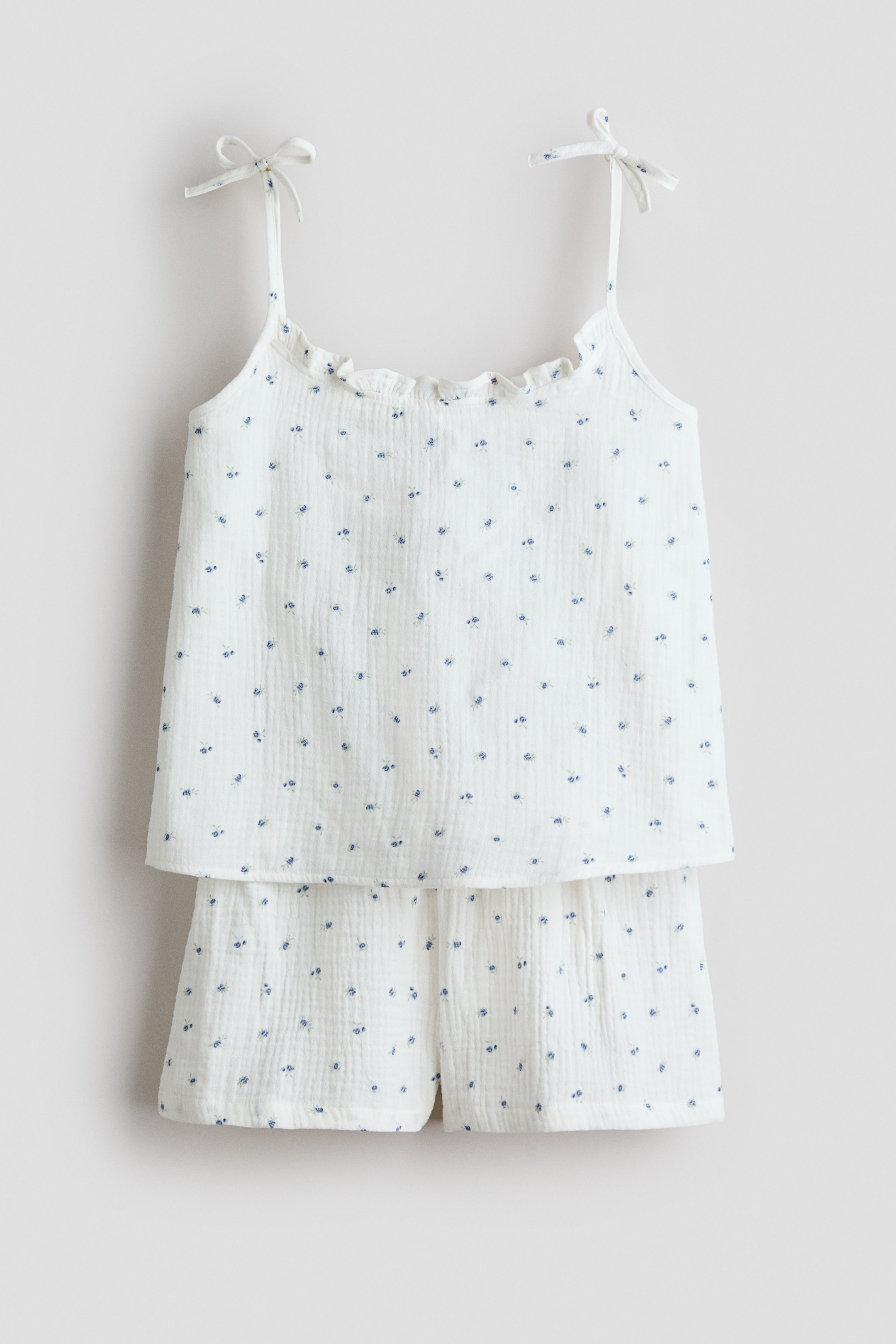 View larger image: Pyjamas featuring a white strappy top and matching shorts, crafted from soft cotton muslin with a delicate blue floral pattern, showcased from the front. The top boasts a frill-trimmed neckline and slender shoulder straps adorned with bows. The shorts display covered elastication at the waist.