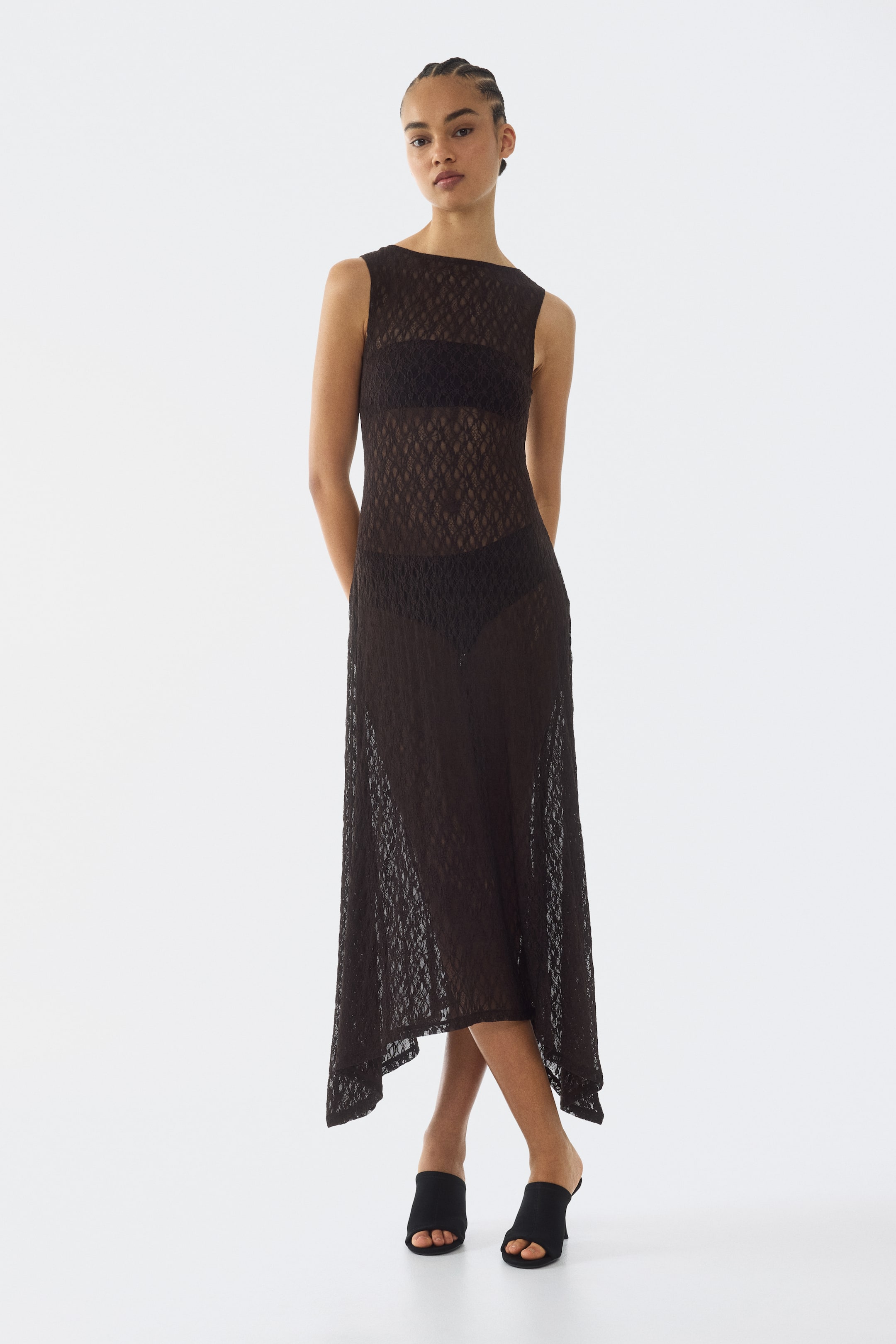 Voir une image plus grande: Une robe maxi noire à motifs transparents est présentée de face, avec un col bateau, un design sans manches et un ourlet asymétrique. Elle est stylée par-dessus un haut bandeau noir et un slip noir, révélant son tissu transparent.