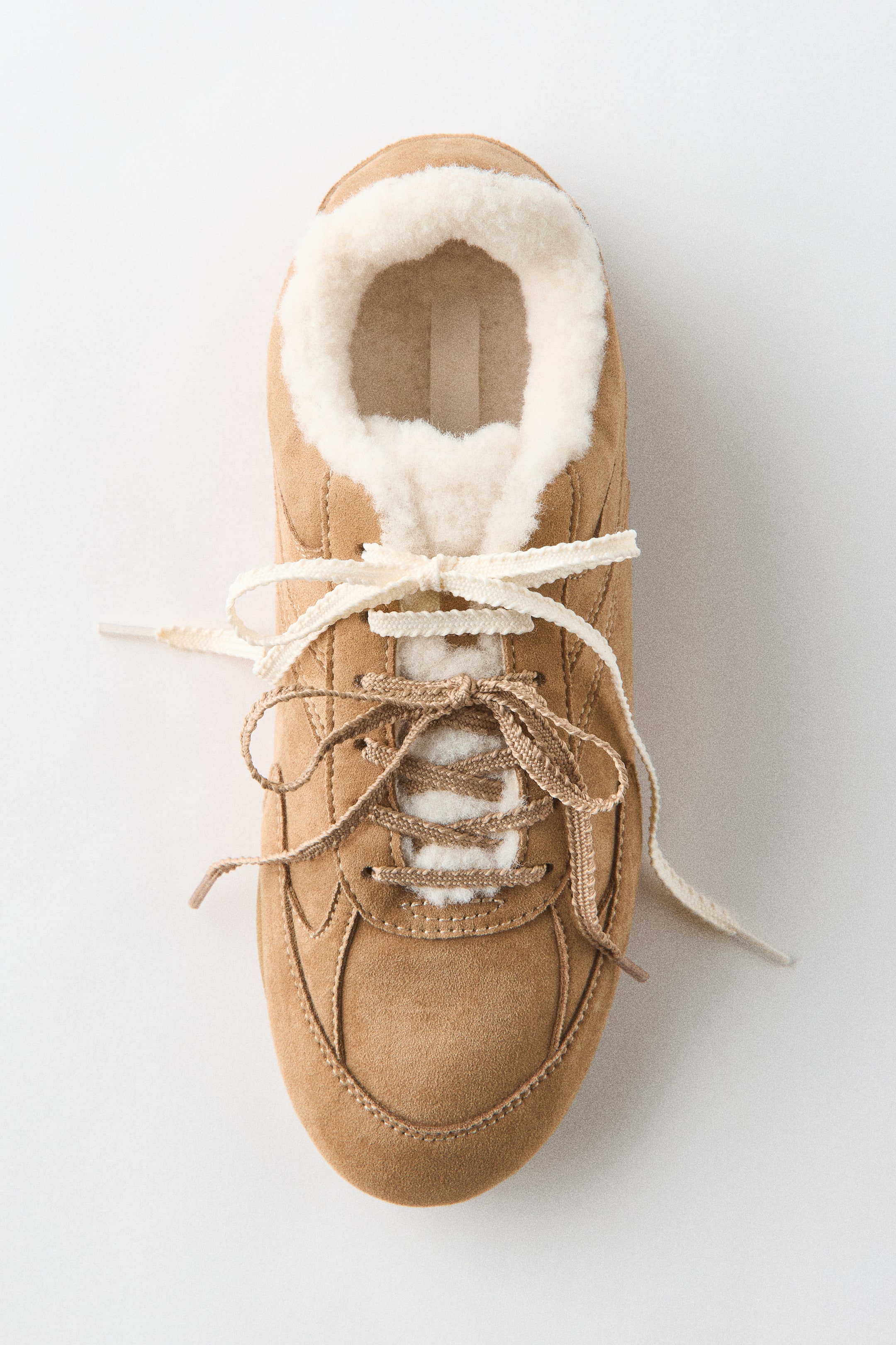 View larger image: A low-profile tan suede trainer is resting in a top-down view facing forwards, showcasing a soft napped finish. The trainer features a fluffy white lining along the collar and tongue, and displays two sets of tied laces in cream and tan.