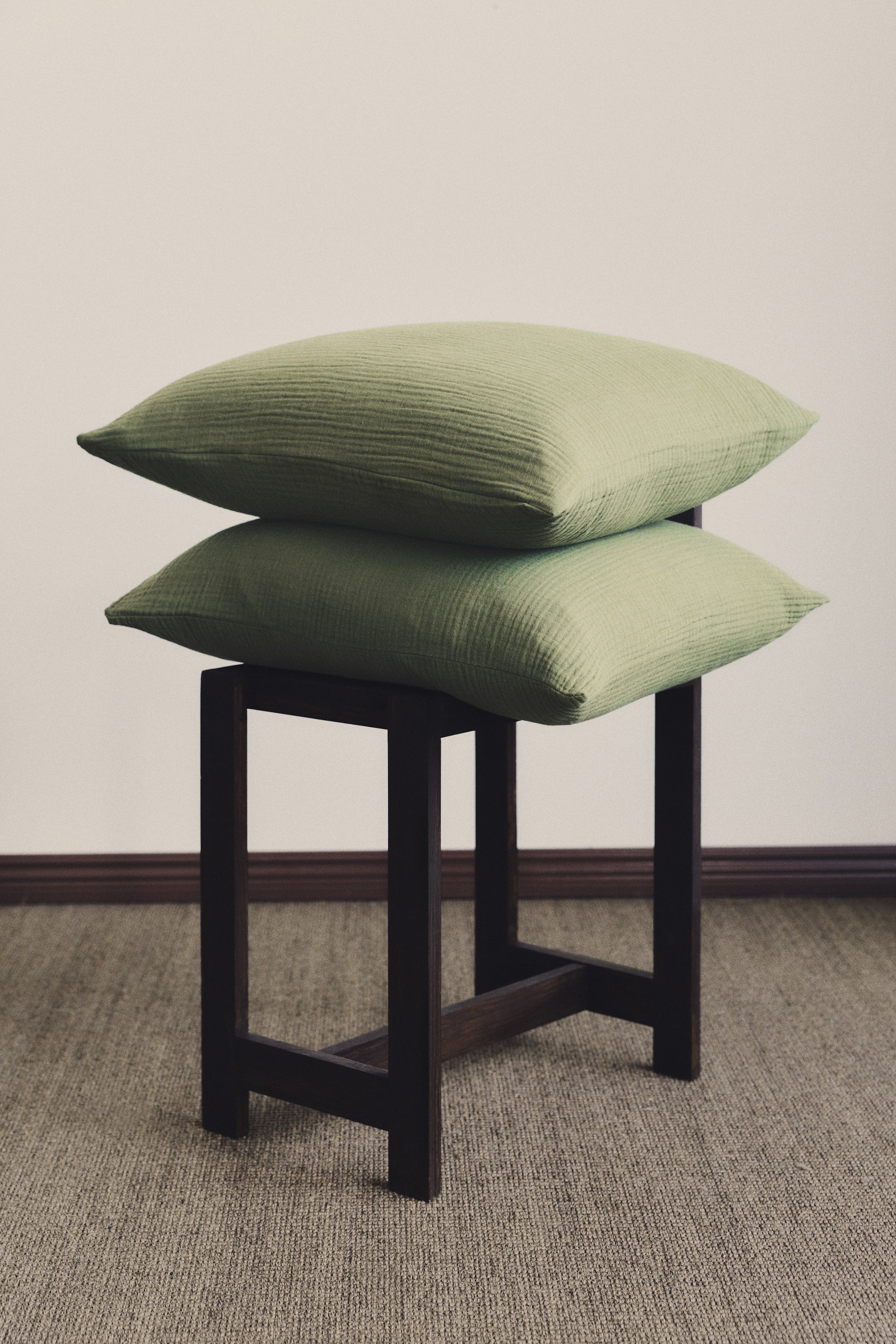 Prikaži veću sliku: Dve jastučnice od mekog pamučnog muslina sa preklopnim otvorom u svetlozelenoj boji i suptilno naboranom teksturom. Postavljene su jedna na drugu, oslonjene na tamnu drvenu stolicu.