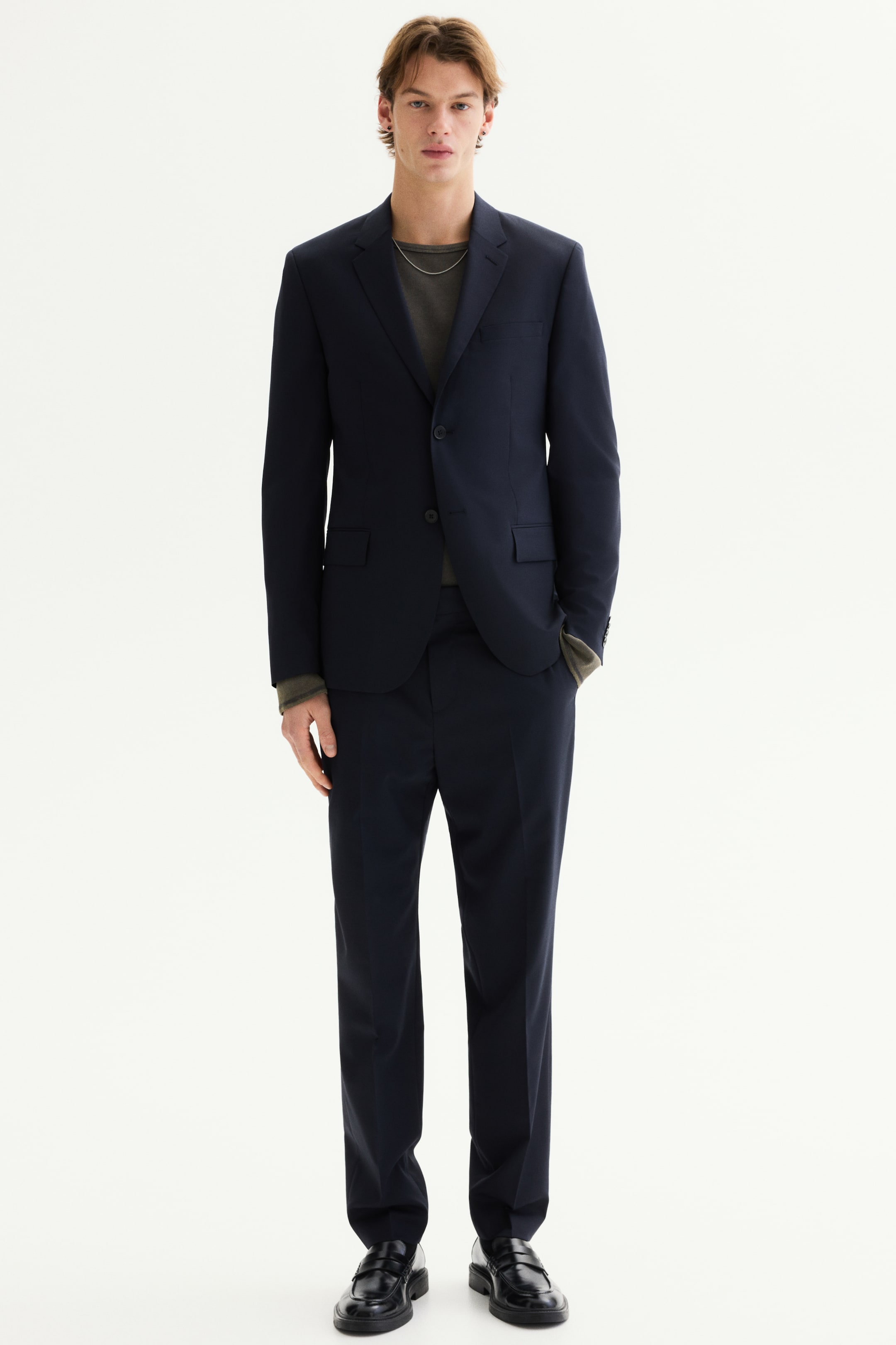 查看大圖: 一套海軍藍西裝正以全長正面視角展示，特色為單排扣西裝外套，帶有缺角翻領、兩顆扣設計和翻蓋口袋，搭配直筒褲。西裝外套搭配灰色圓領上衣，褲子則搭配黑色樂福鞋。