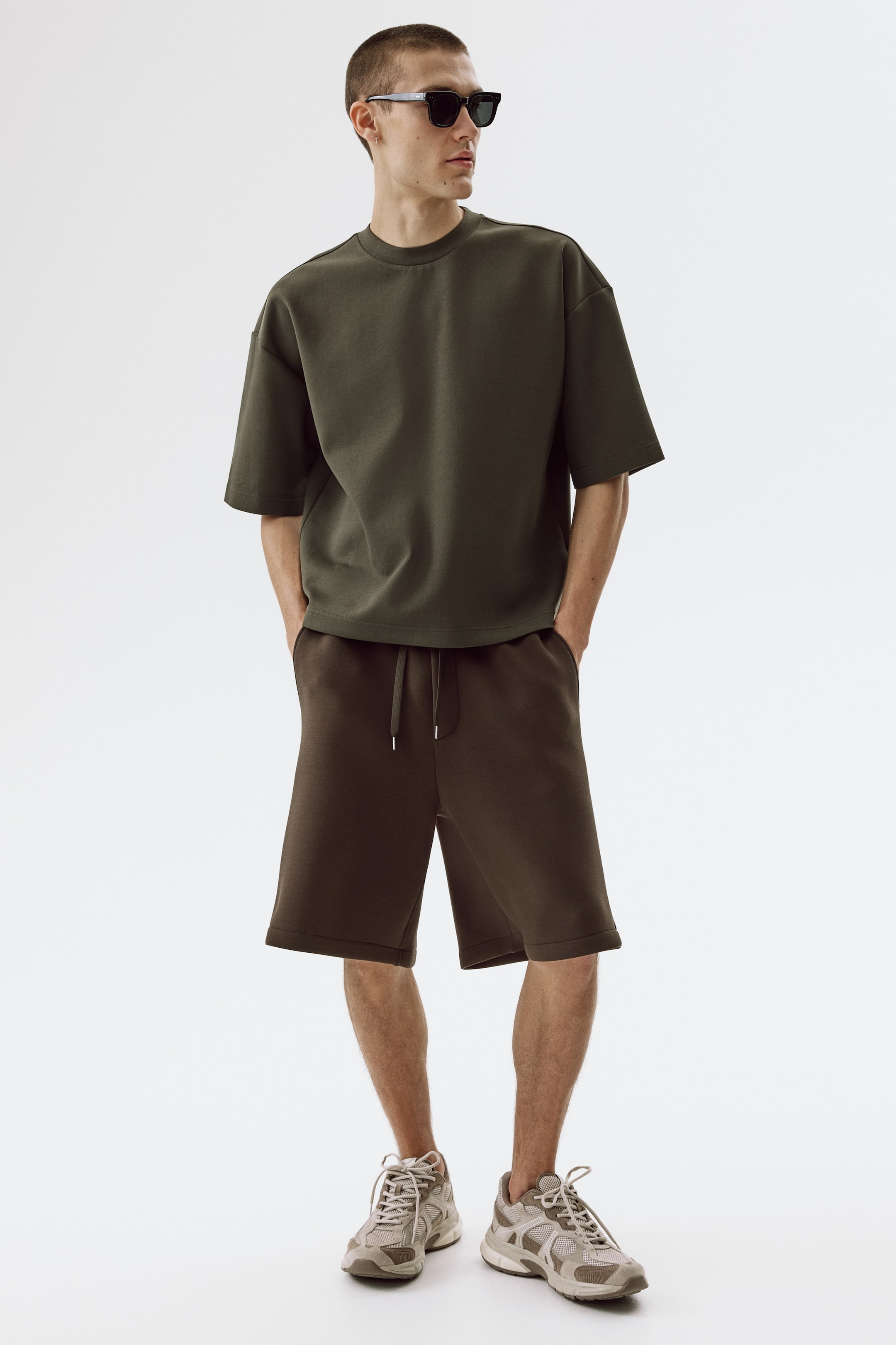 Vis større bilde: Den mørke olivengrønne oversized t-skjorten med korte ermer og rund hals bæres, og komplementerer de mørkebrune knelange shortsene med elastisk snøring i midjen. Begge plaggene har en avslappet passform og en glatt, tykk stofftekstur.