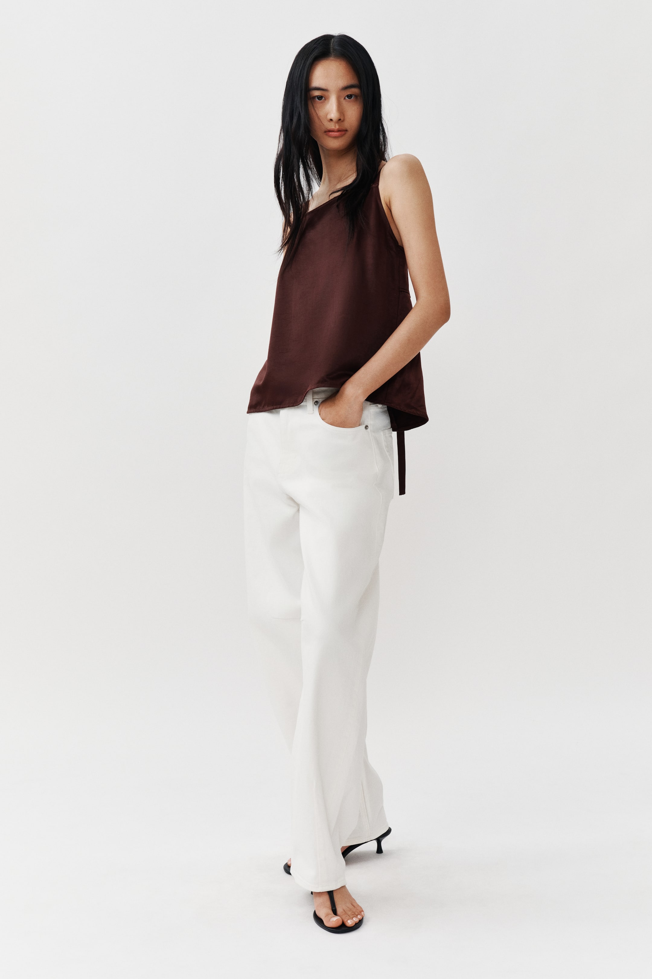 View larger image: A dark brown, satin-like one-shoulder top featuring an asymmetric neckline and a loose fit. It is styled with white wide-leg pants, partially tucked in on one side, and black thong-style heeled sandals.