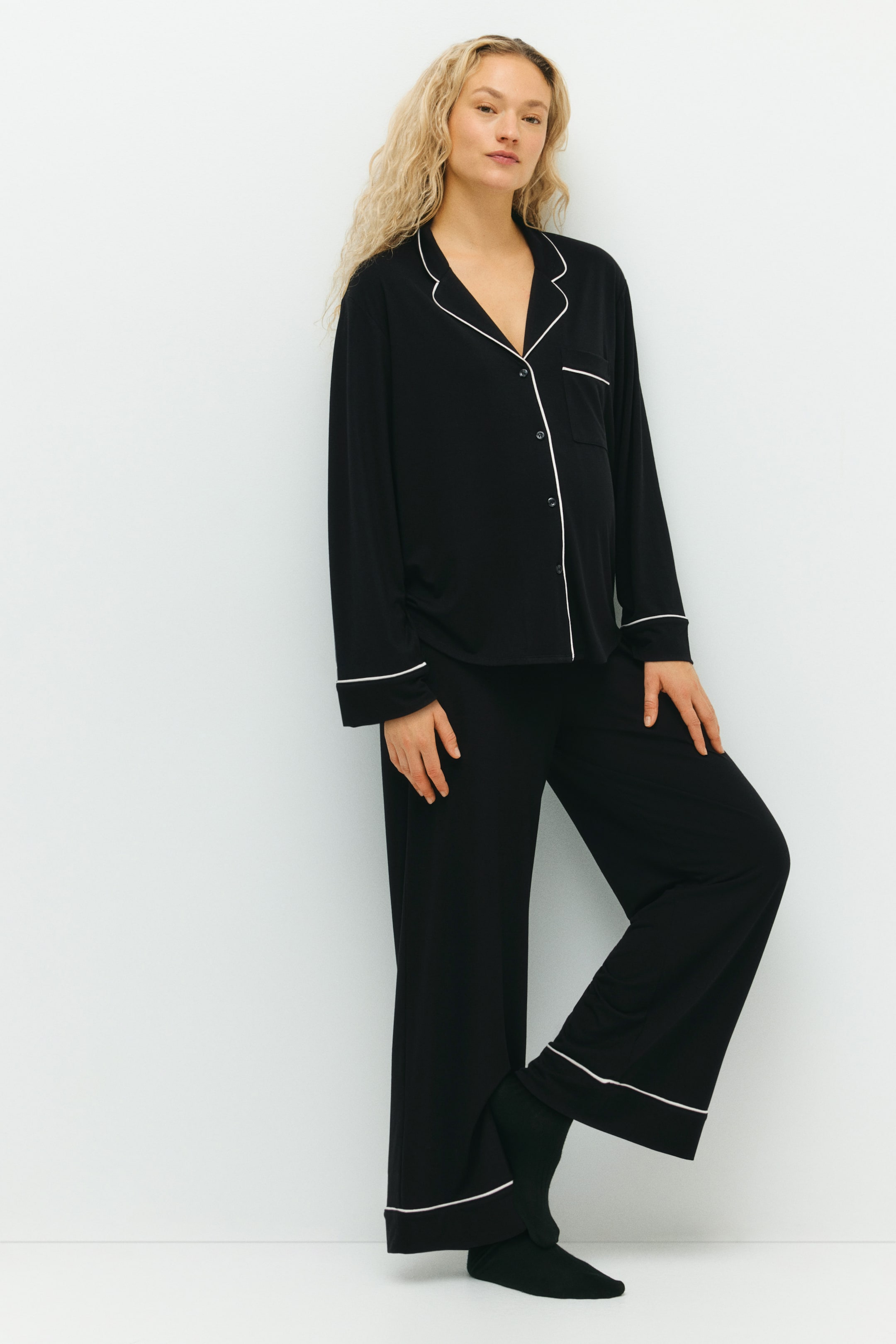 Größeres Bild ansehen: Schwarze Pyjamas aus weichem Jersey, bestehend aus einem Hemd mit Resort-Kragen, Frontknopfleiste, einer offenen Brusttasche und langen Ärmeln, sowie weiten Beinabschlüssen, werden von dem Model getragen. Zarte weiße Paspeln akzentuieren den Kragen, die Knopfleiste, die Manschetten und die unteren Säume des Hemdes, während das Model mit einem angewinkelten Bein steht.