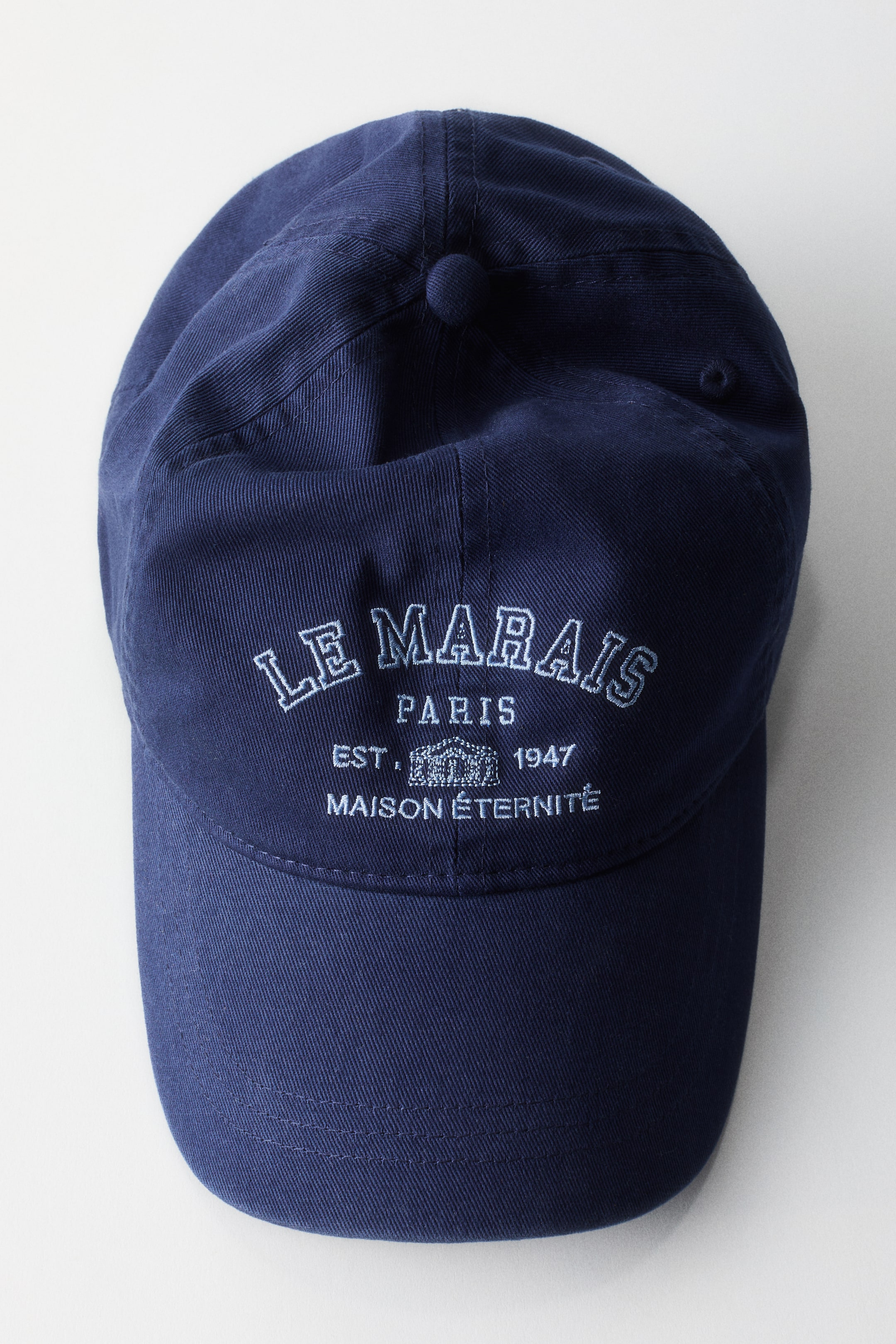 Se større billede: En marineblå baseballkasket med en seks-panels konstruktion og en buet skygge. Frontpanelet viser hvidt broderet tekst, der lyder "LE MARAIS PARIS EST. 1947 MAISON ÉTERNITÉ".