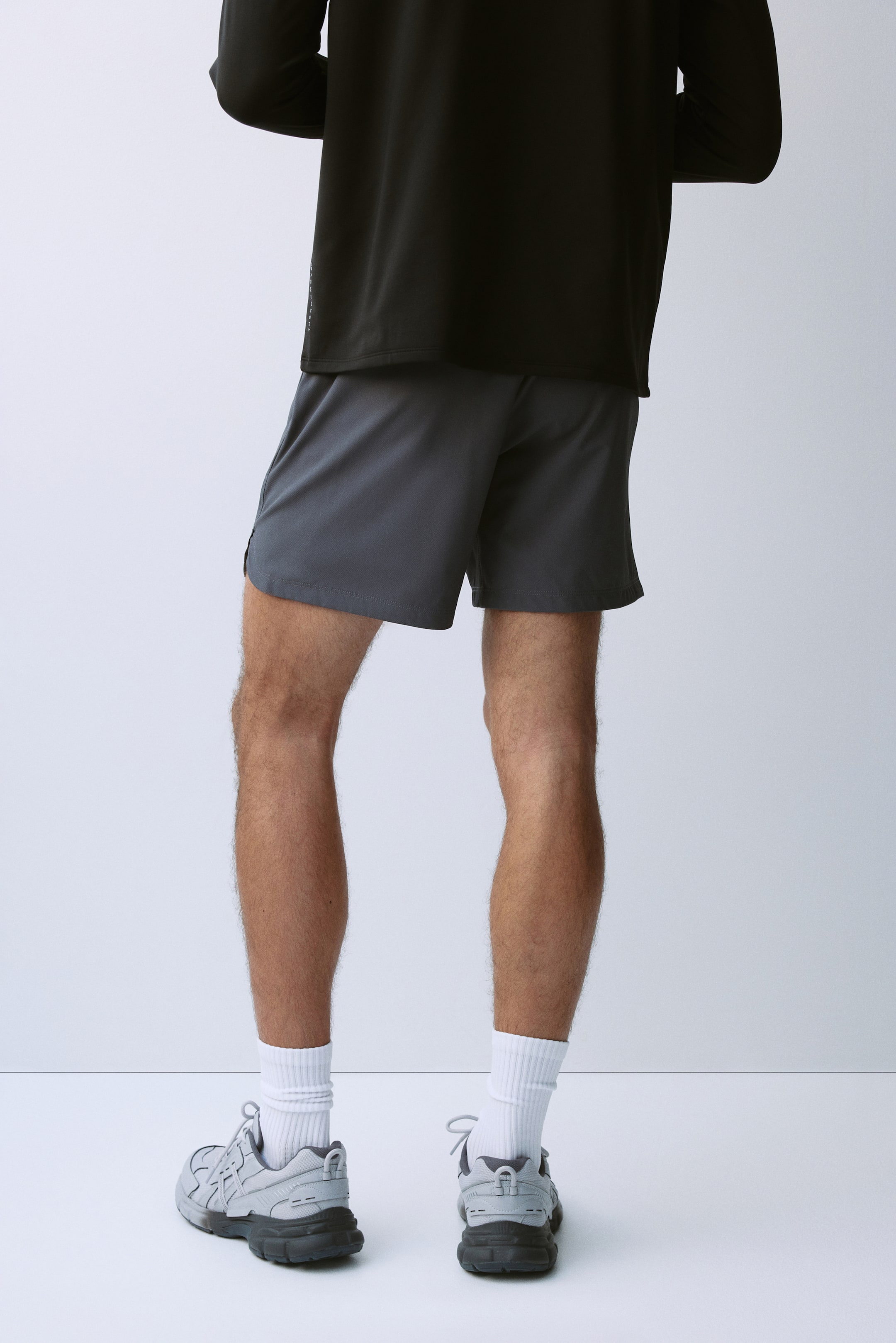 View larger image: Grey athletic shorts are being worn by the model, shown from a rear view. The mid-thigh length shorts are styled with a black long-sleeve shirt, white ribbed crew socks, and grey athletic shoes.