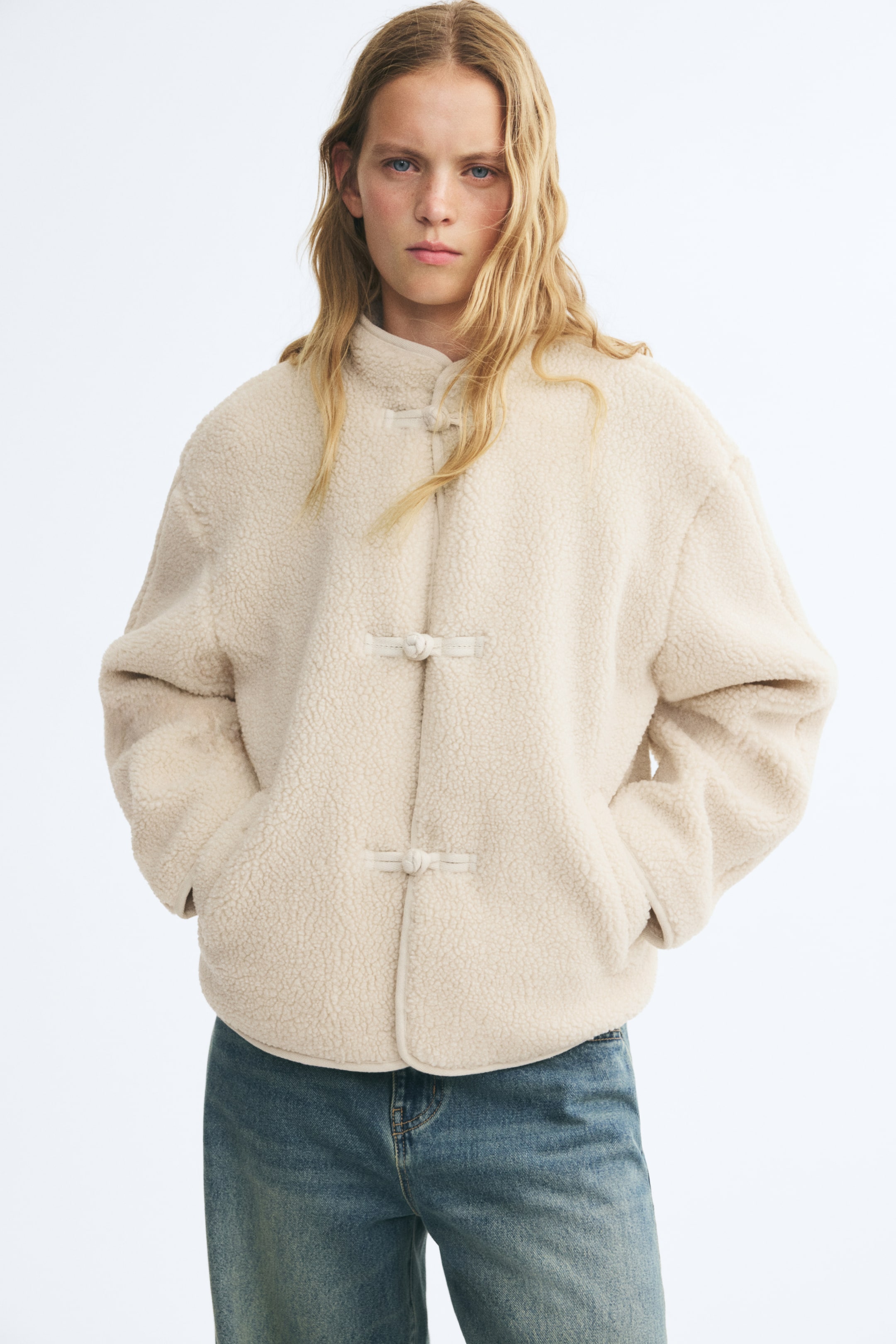View larger image: A cream-colored, relaxed-fit teddy jacket with a stand collar, dropped shoulders, and front knot buttons is worn by the model. The model is standing with hands placed in the jacket's welt pockets, paired with blue denim jeans.