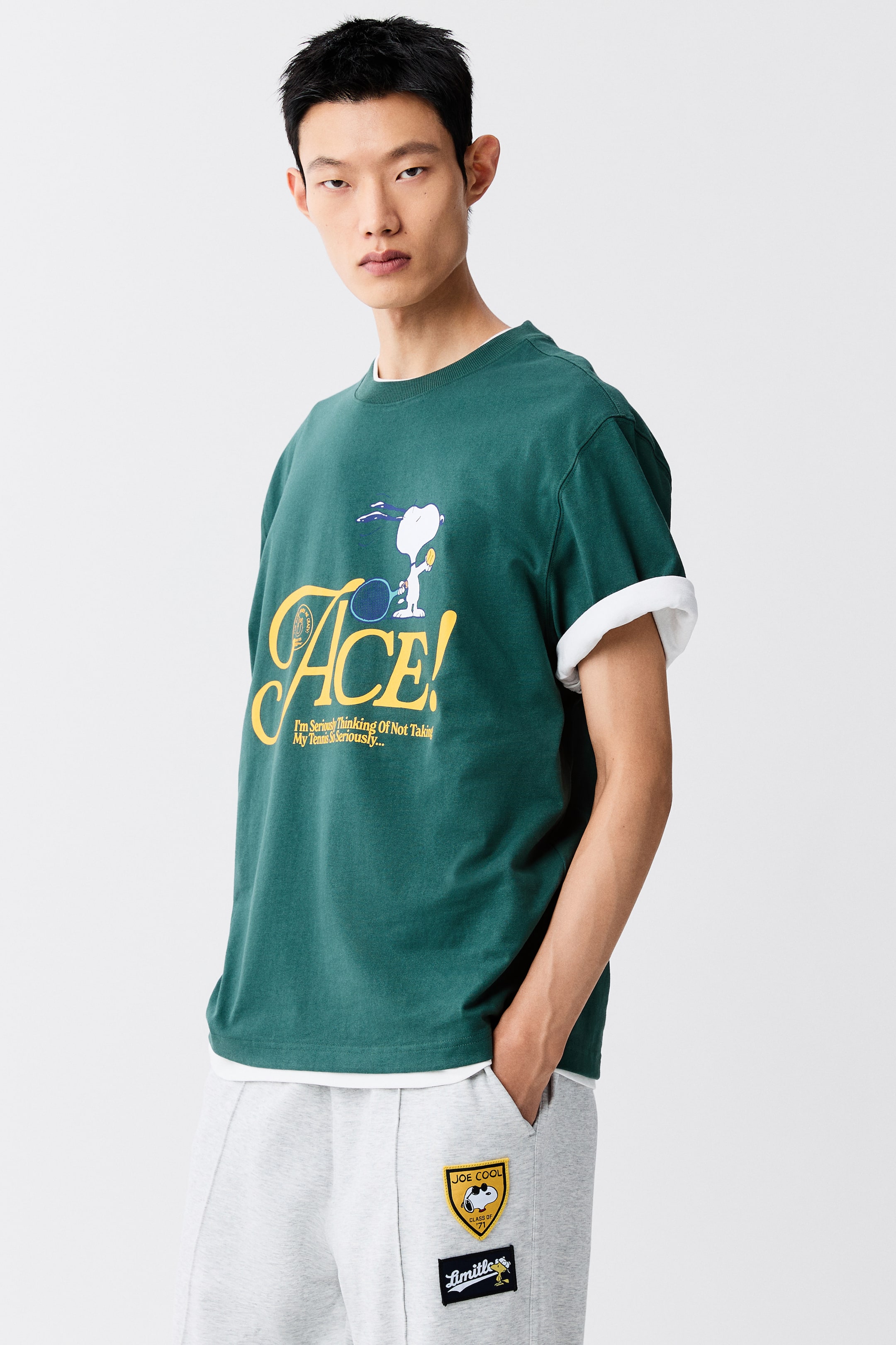 View larger image: A dark green heavyweight cotton t-shirt featuring a printed Snoopy graphic is worn by the model. The loose-fit tee is layered over a white t-shirt with rolled-up sleeves and styled with light grey sweatpants.