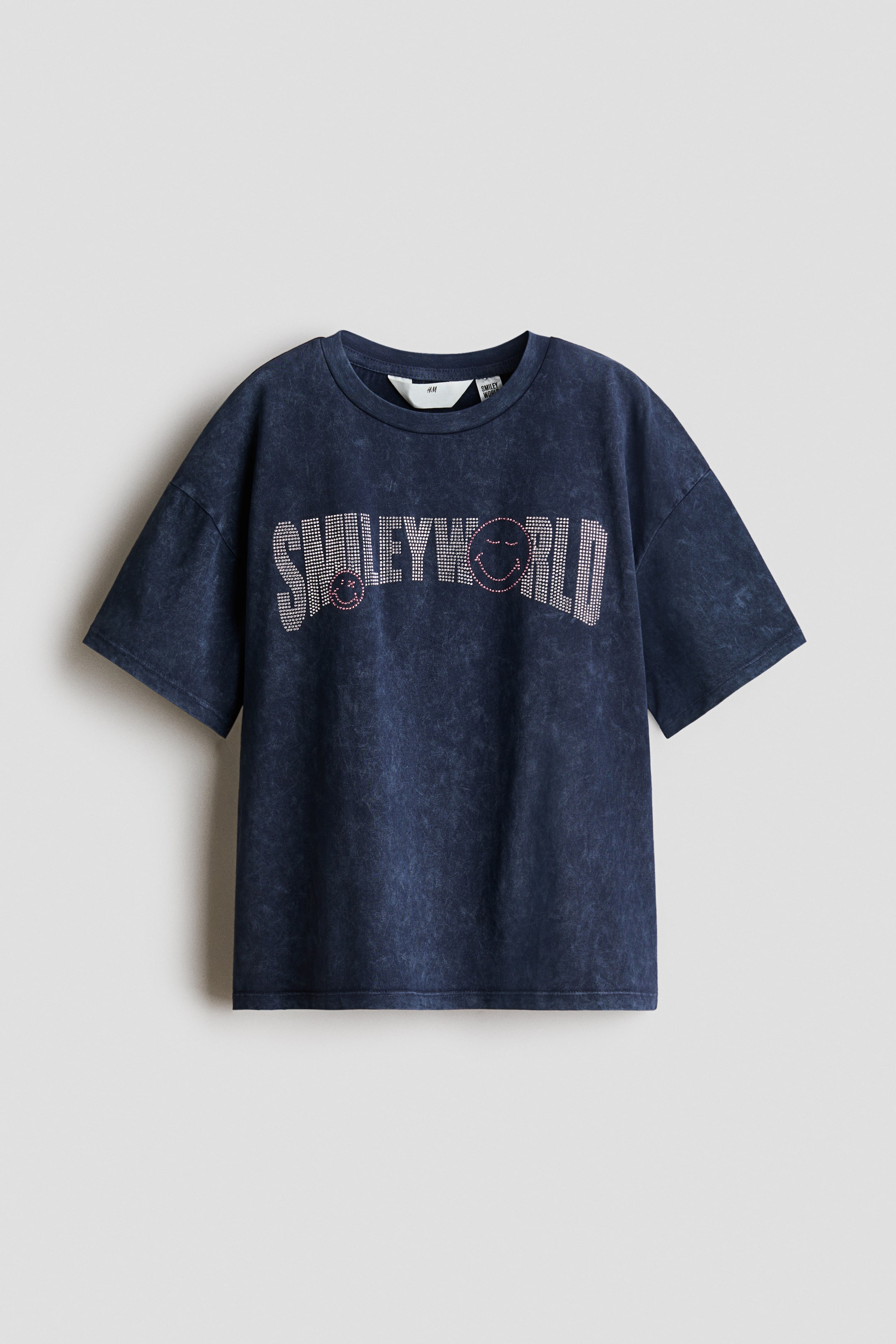 Agrandir l'image: Un t-shirt bleu foncé délavé en jersey de coton doux est présenté de face, arborant un grand imprimé "SMILEYWORLD" sur la poitrine. Le t-shirt est conçu avec un col rond bordé de côtes et des épaules tombantes, et l'imprimé est composé d'éléments brillants formant les lettres et deux visages souriants aux yeux fermés avec des accents roses.