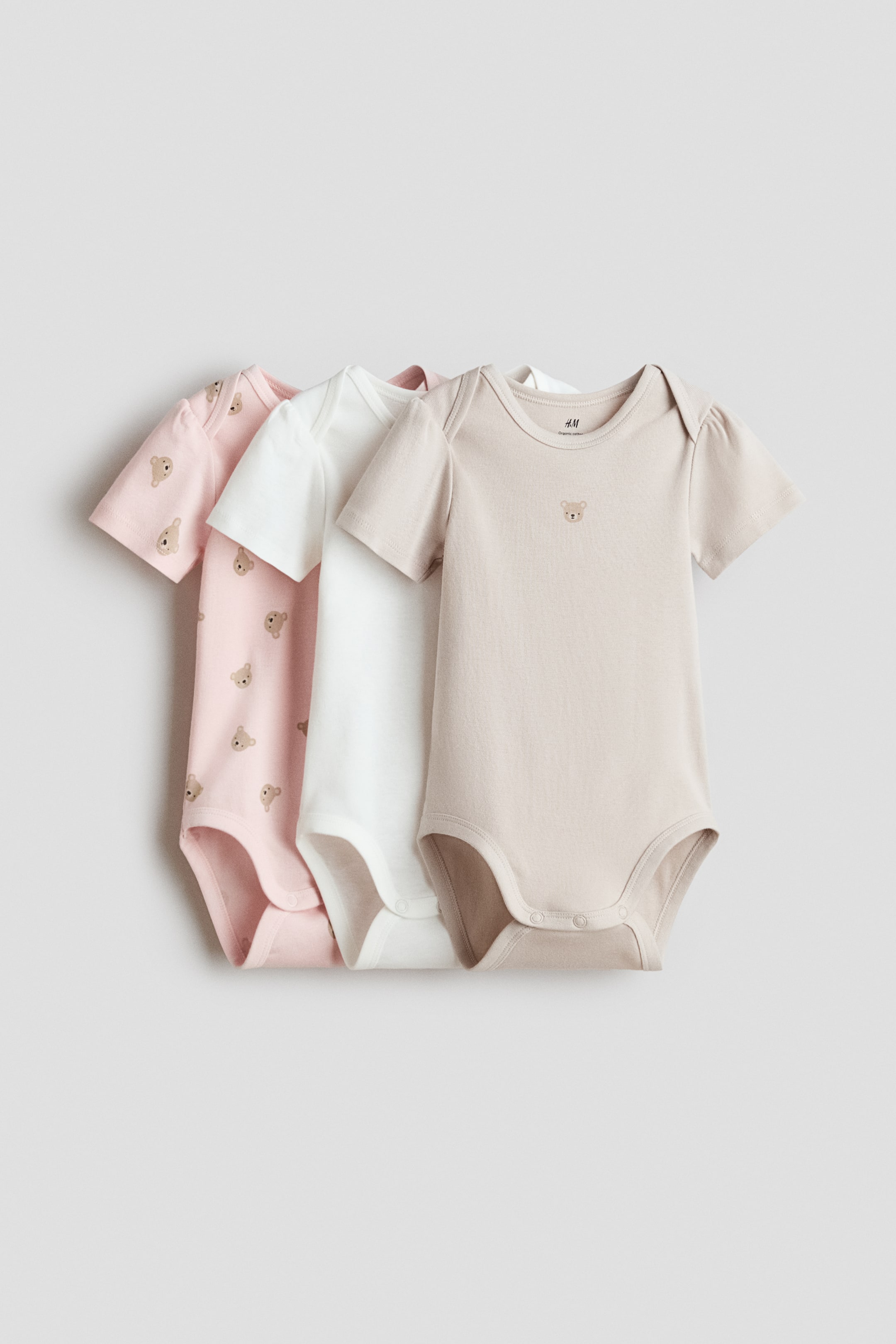 View larger image: Three short-sleeved bodysuits are showcasing their fronts, featuring an envelope neckline, snap buttons at the bottom, and gathered leg openings. The pink bodysuit displays a repeating bear head pattern, the white one is solid, and the beige one features a small embroidered bear head on the chest.